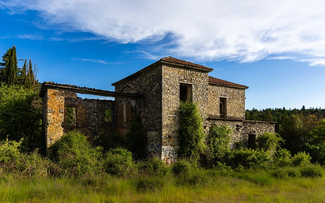 Old abandoned stone house surrounded by overgrown vegetation under a blue sky with clouds.