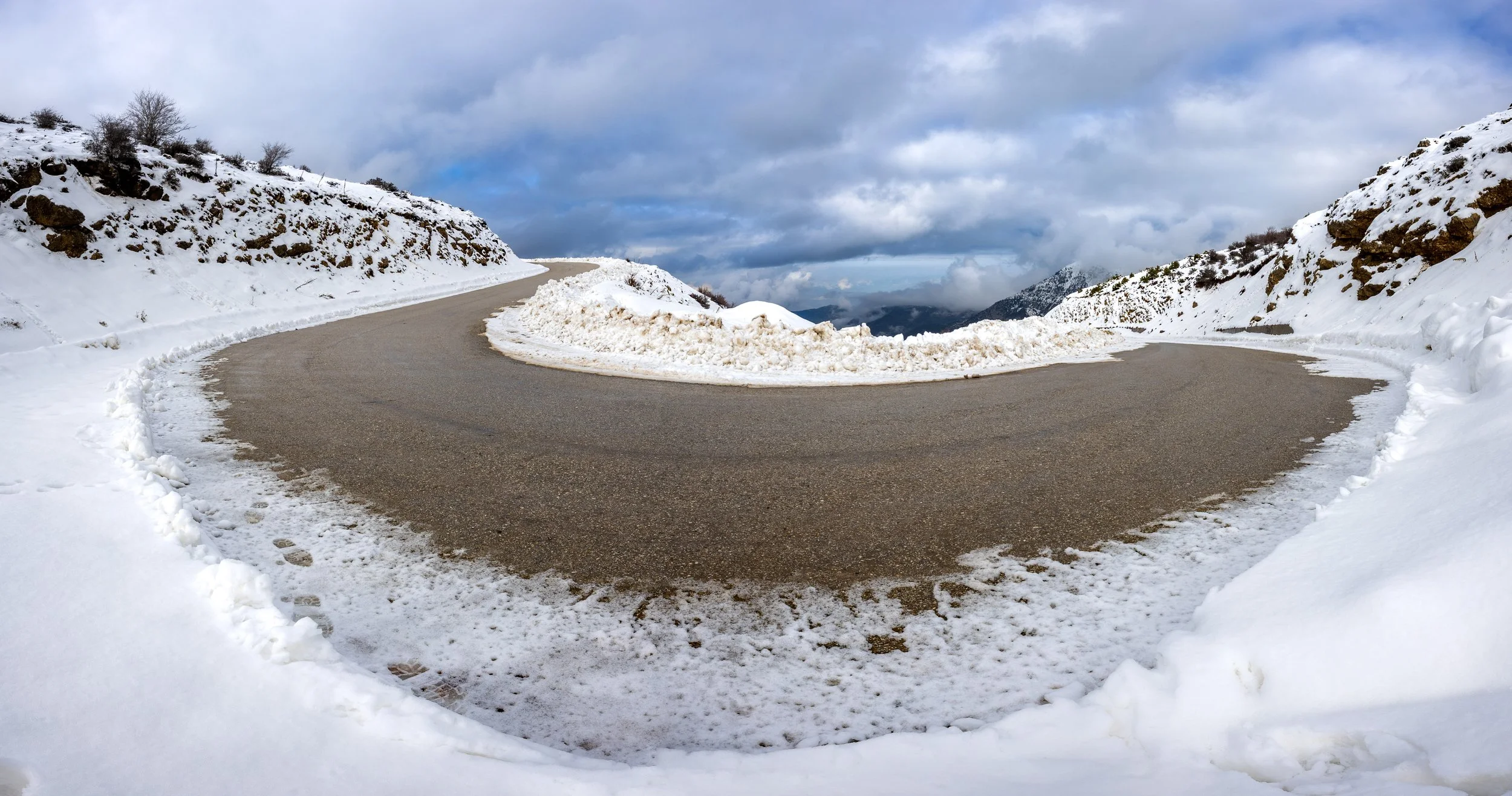 A winding mountain road with snow on the sides and a snowy landscape, with cloudy sky overhead photography by erez nudmanov