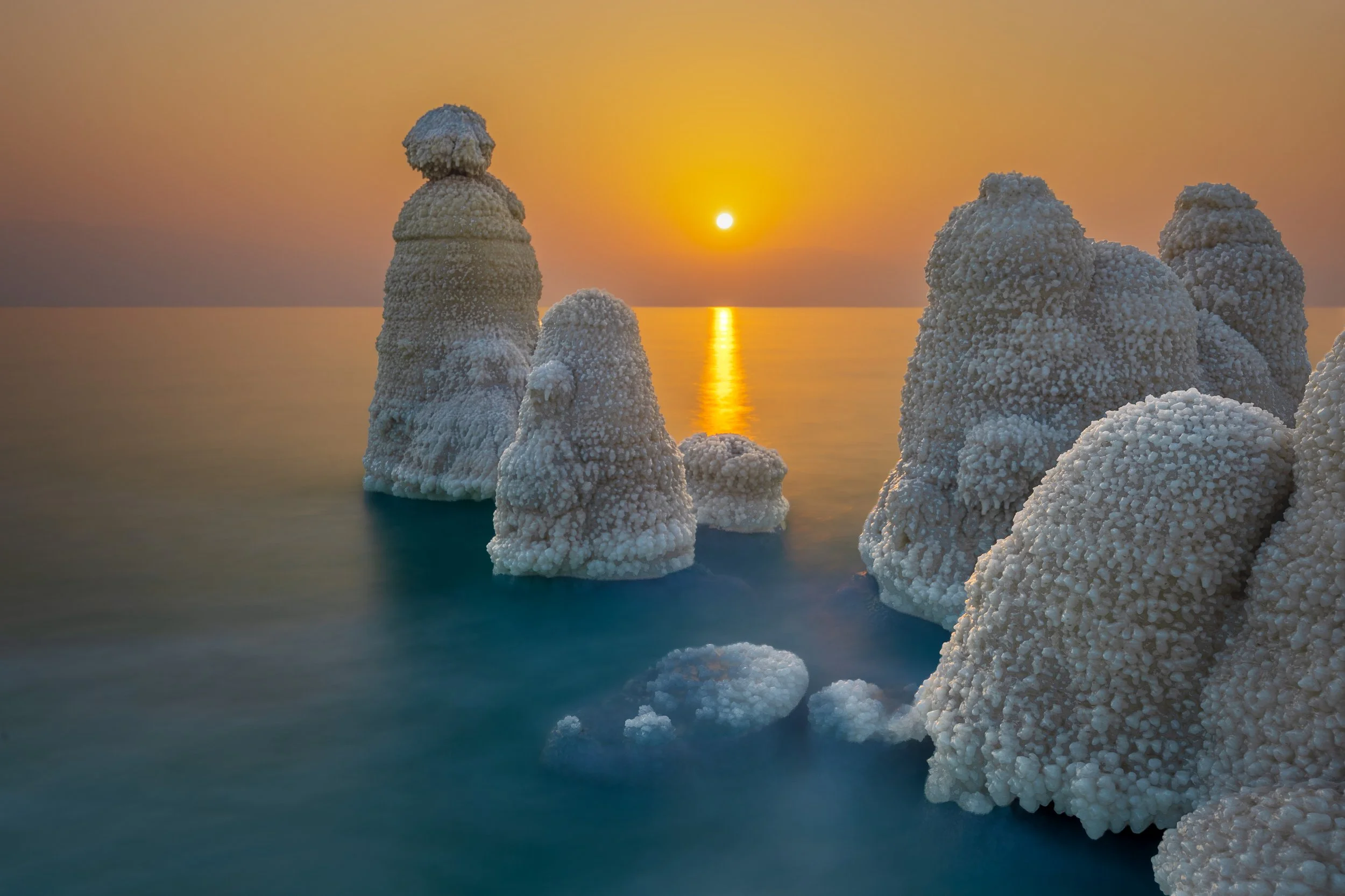 Salt formations resembling rocks along a calm sea at sunset, with the sun low on the horizon casting a golden reflection on the water.