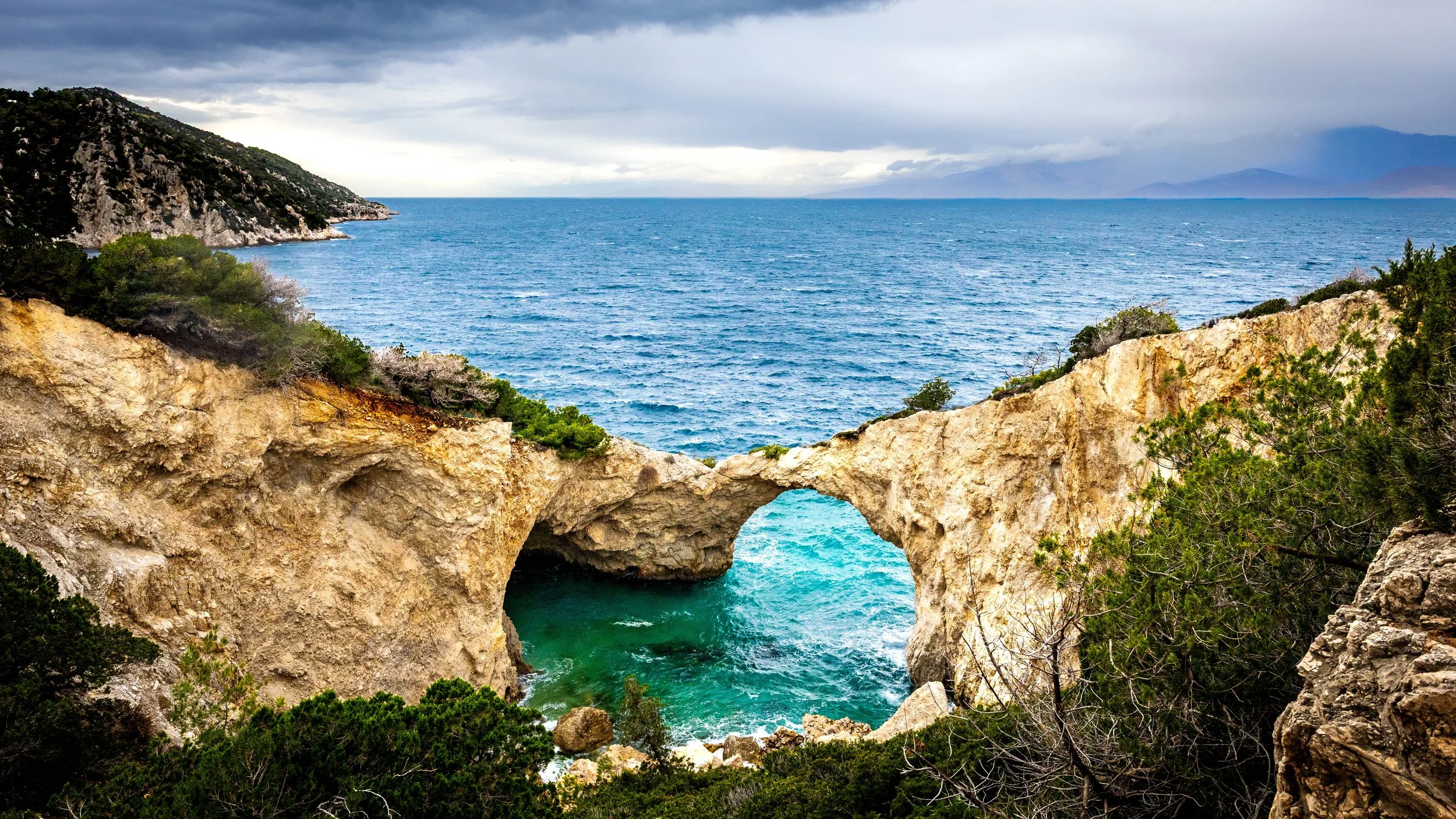 A coastal view of a natural rock arch formation with the ocean beyond, surrounded by green shrubs and mountains in the distance under cloudy skies, photograph by erez nudmanov