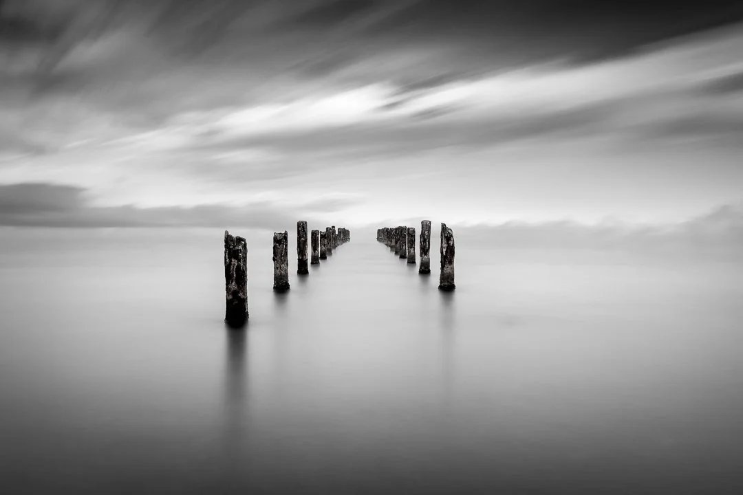 Black and white long exposure photo of old, weathered wooden posts in calm water, with cloudy sky above.