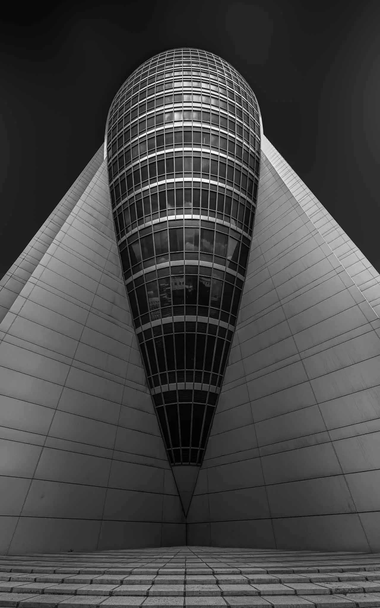 Black and white architectural photograph of Haifa’s Sail Tower, viewed symmetrically from below between converging geometric walls.