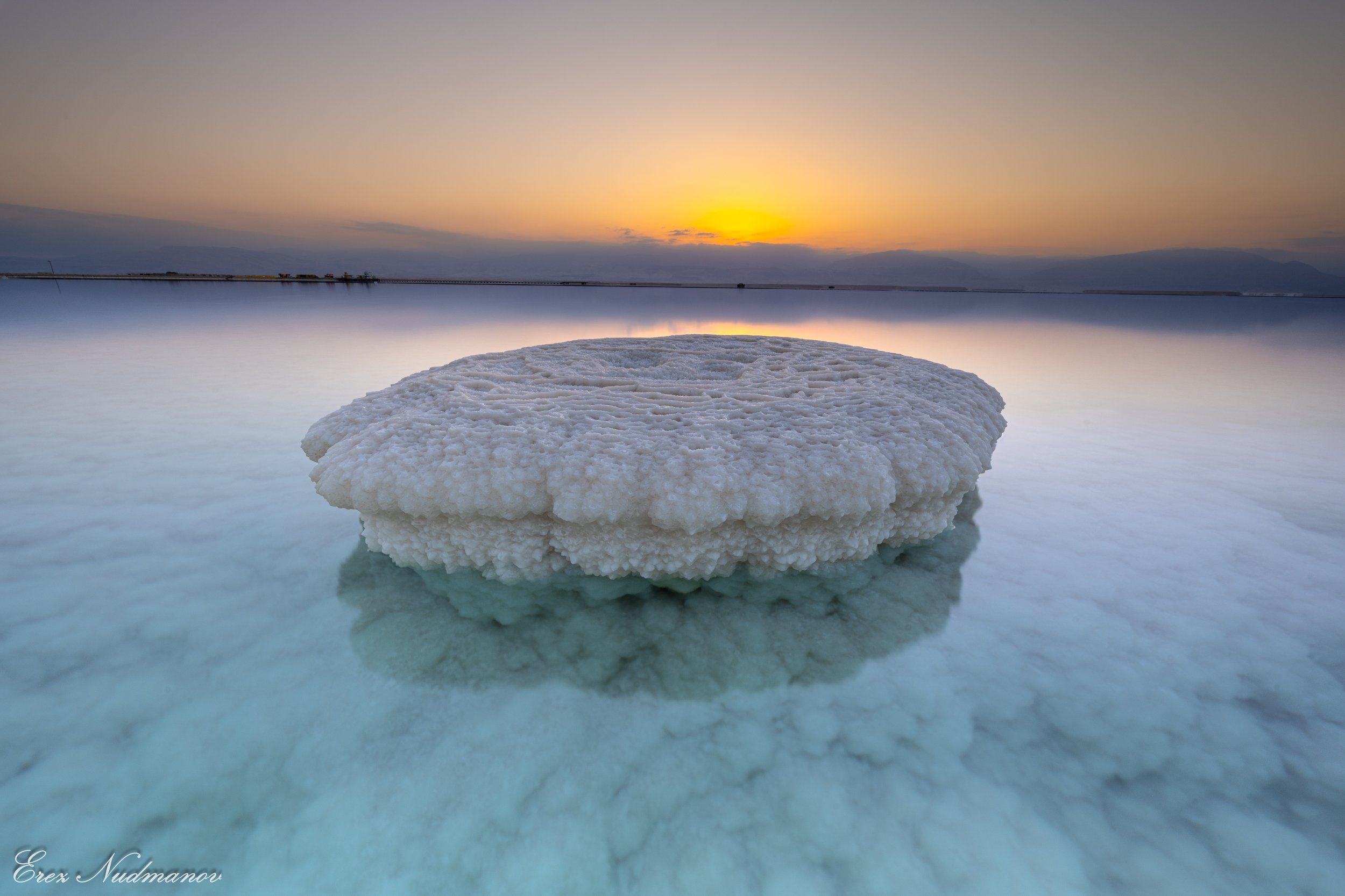 A salt formation floating on a calm body of water at sunset, with the sky showing a gradient of colors from orange to purple.
