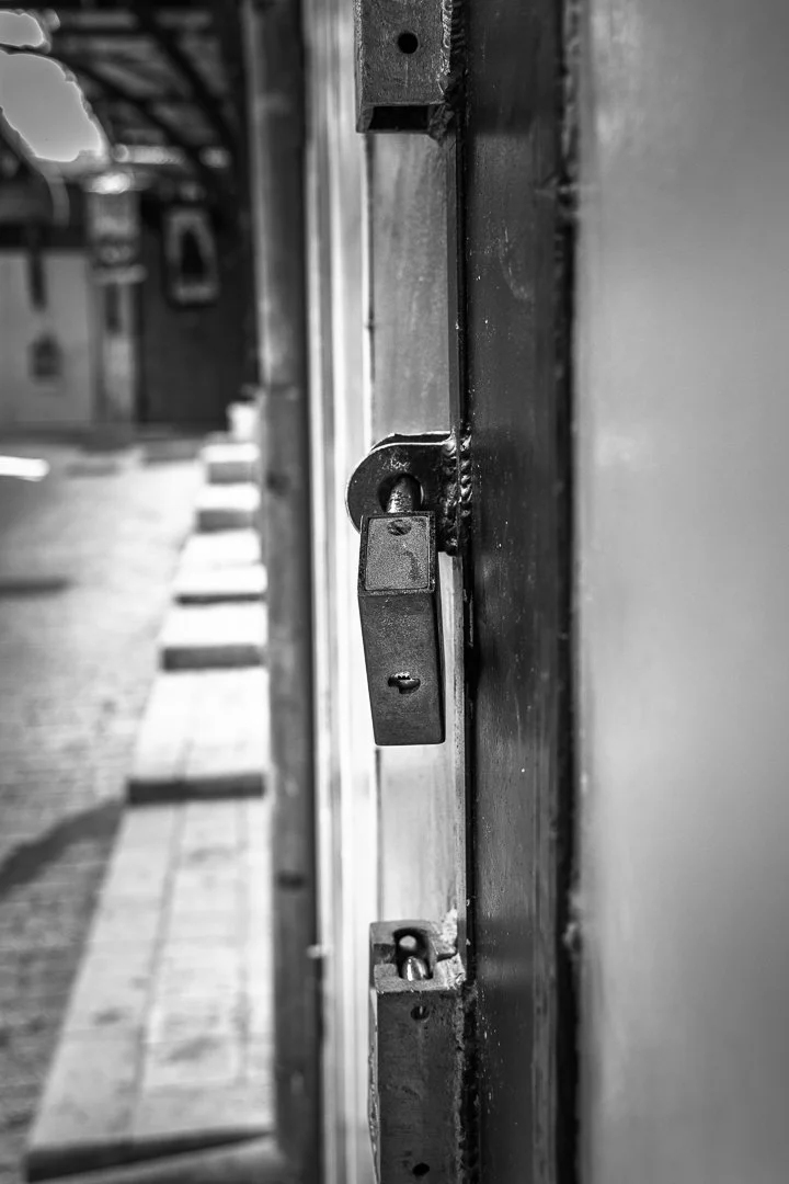 Close-up of a locked door lock on a metal door, with a blurred sidewalk and building in the background.