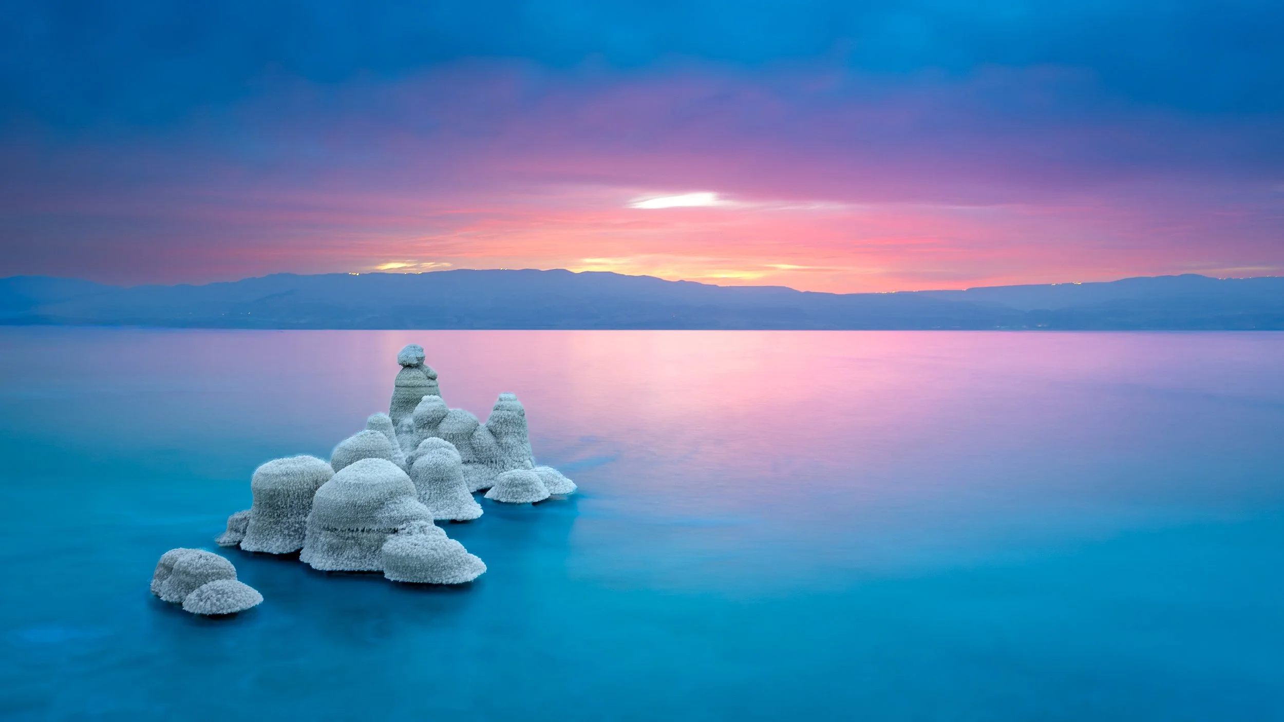 Frost-covered rock formations in a calm lake at sunset with colorful sky and distant mountains.