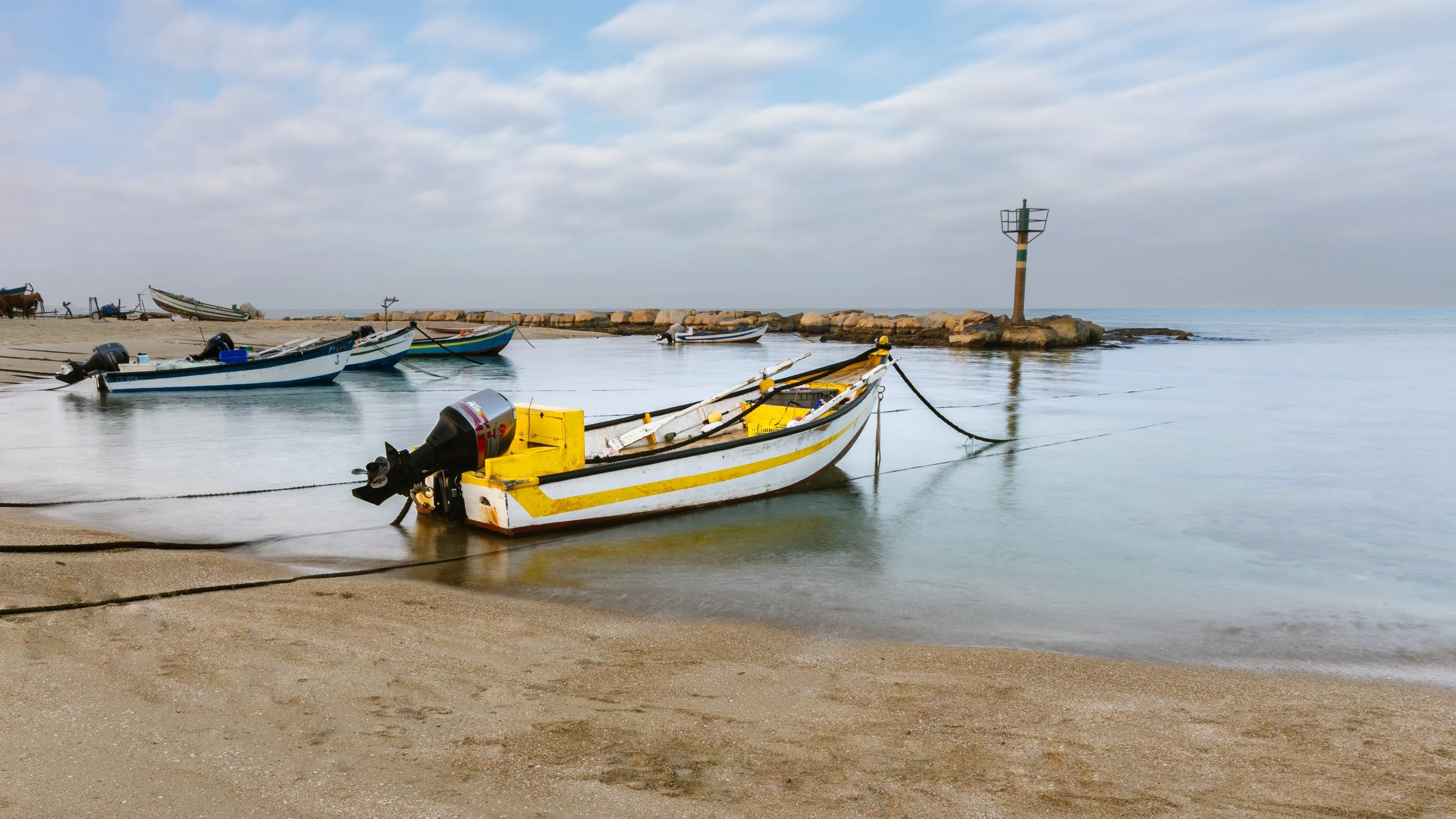Small yellow and white motorboat docked near the sandy shore of a calm beach, with several other boats moored in the background and a rocky jetty with a navigational marker extending into the water.