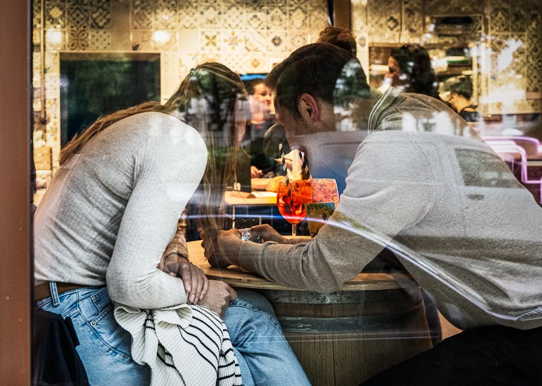 A man and a woman are sitting at a table inside a restaurant, engaged in an arm wrestling match, viewed through a window with reflections of the street outside.