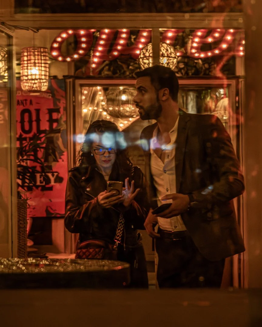 A man and woman stand inside a decorated venue, seen through a glass window at night. The woman is looking at her phone, wearing glasses and a leather jacket. The man, taller and in a suit jacket, looks to the side. Behind them, warm lights and a sig