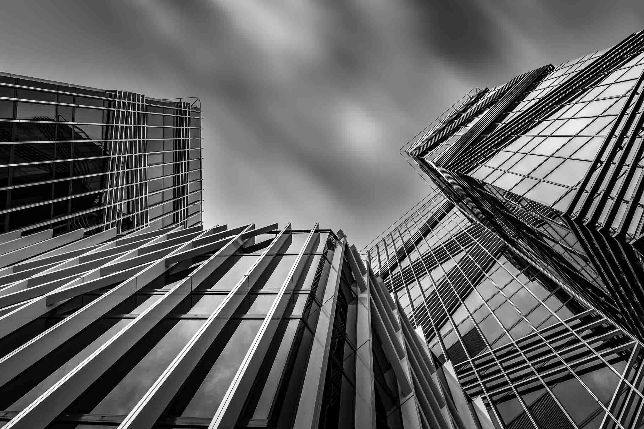 Black and white fine art architectural photograph looking upward between modern glass buildings, with converging geometric lines and motion-blurred clouds.