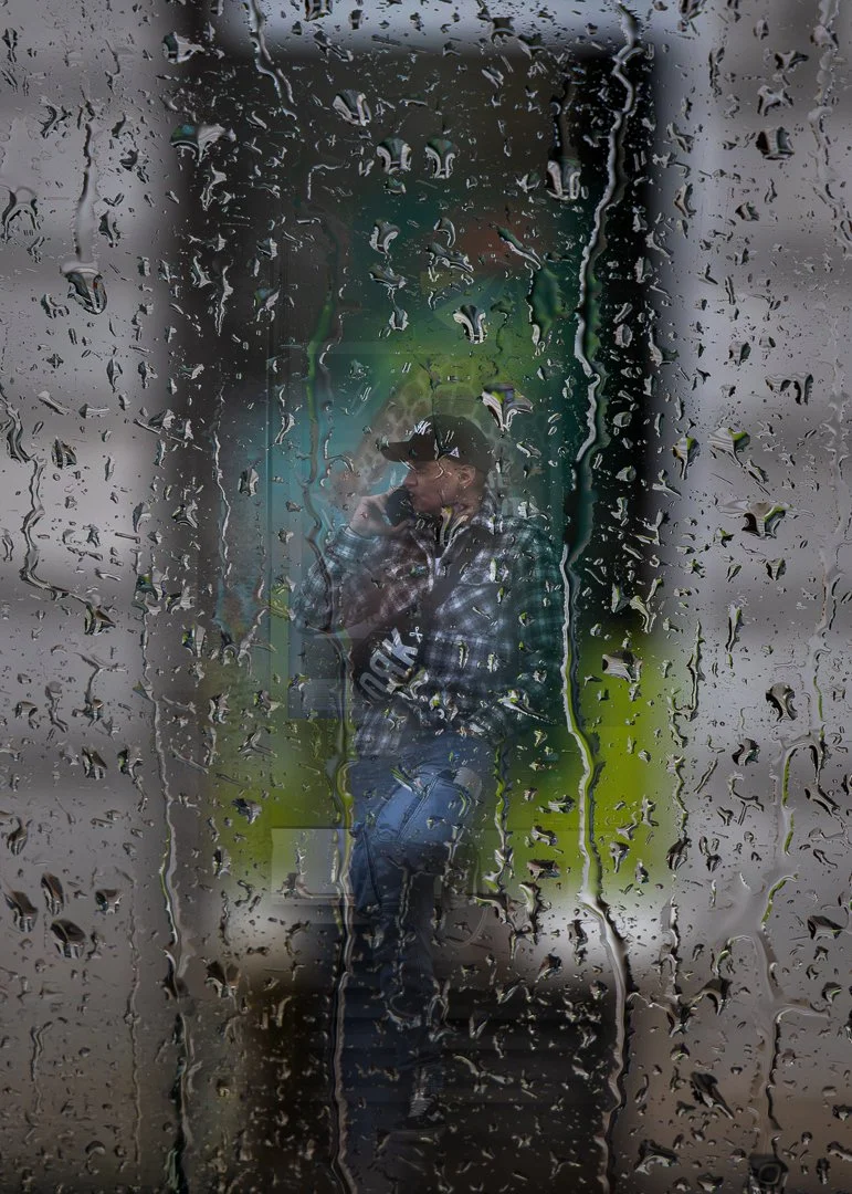 A person talking on a cell phone, reflected in a window covered with raindrops.