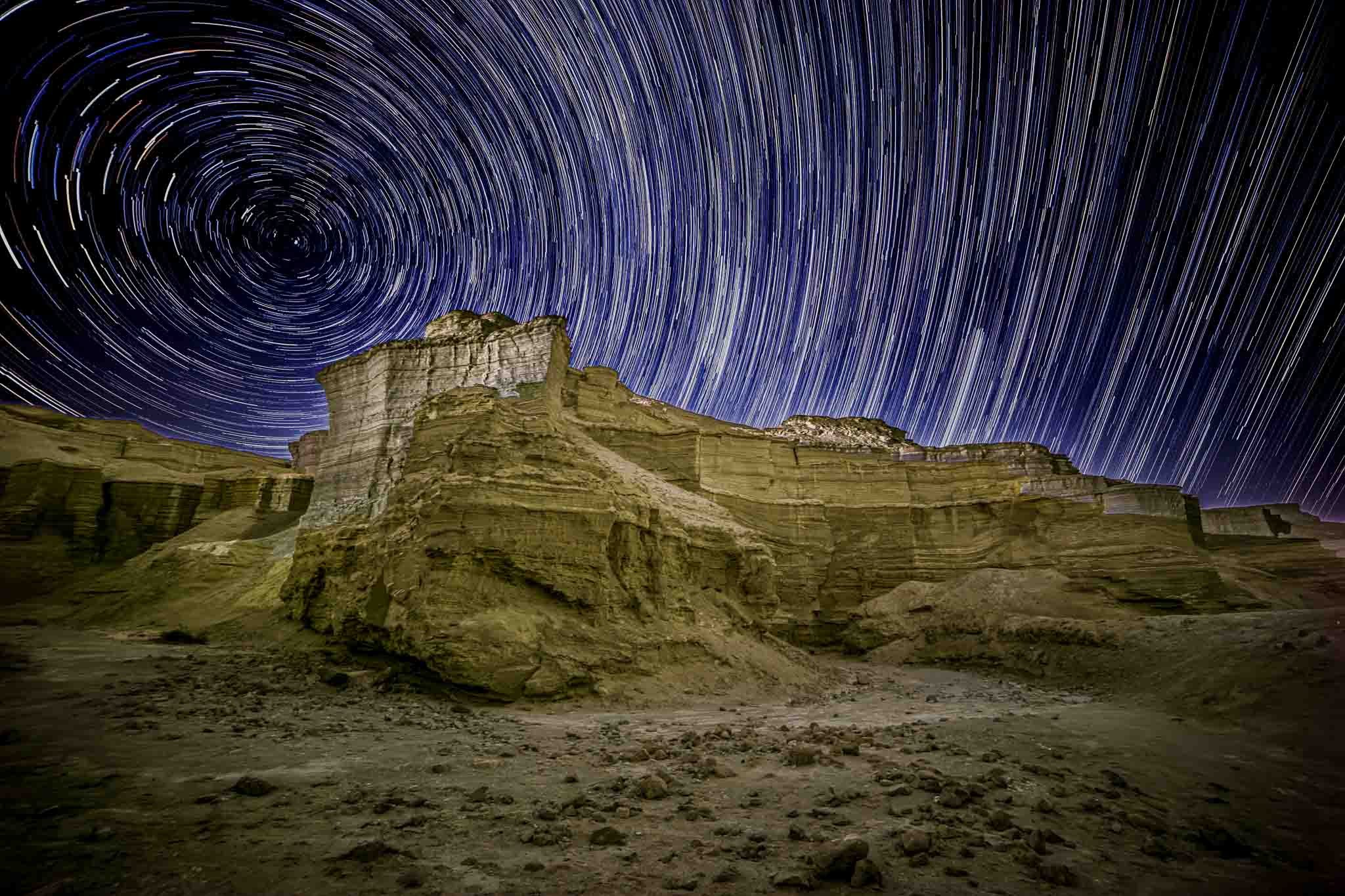 Star trails curve above the Havarei Masada desert formations in a long-exposure night photograph near the Dead Sea.