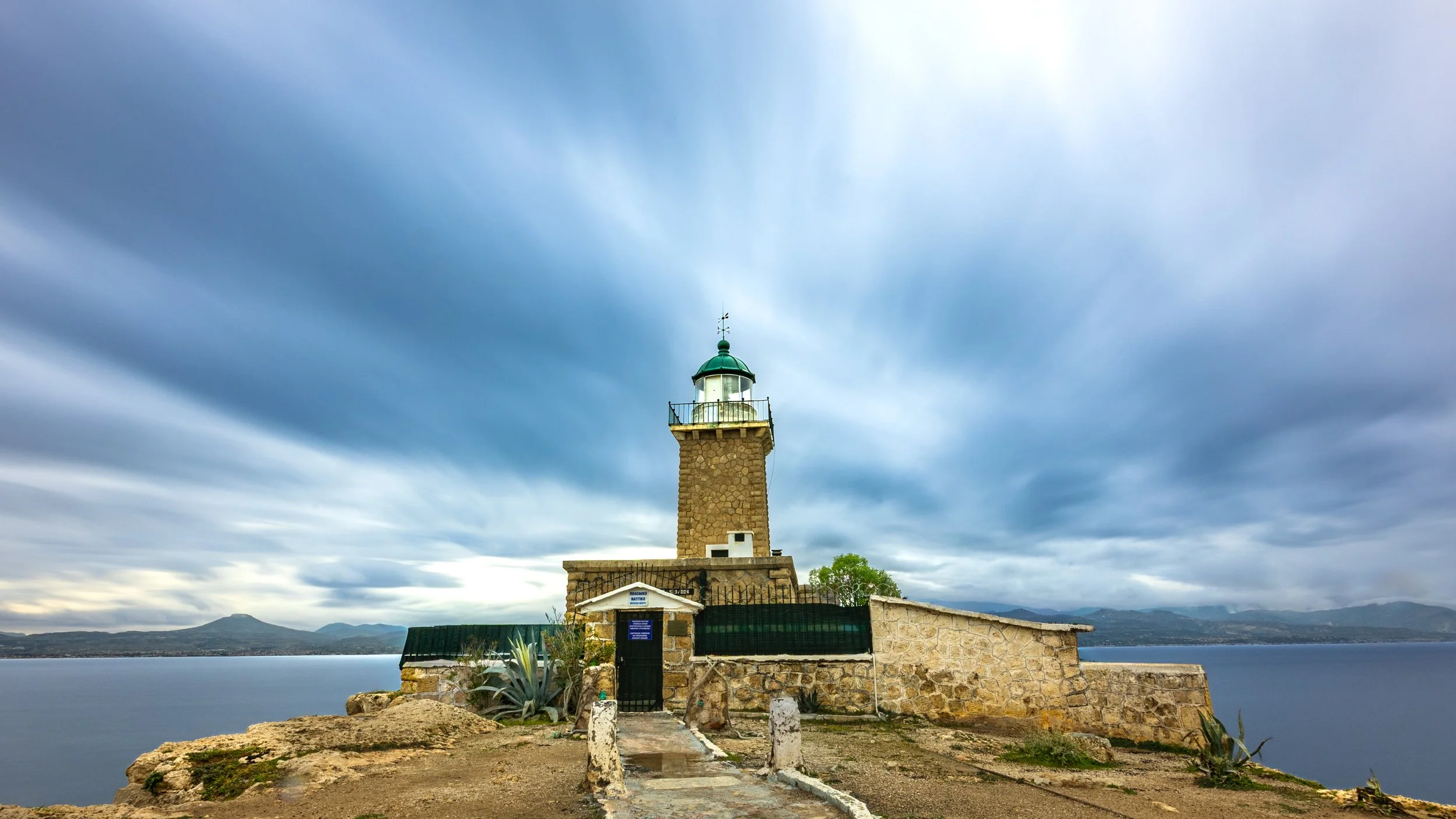 A lighthouse on a rocky coast with water and mountains in the background, under a cloudy sky.