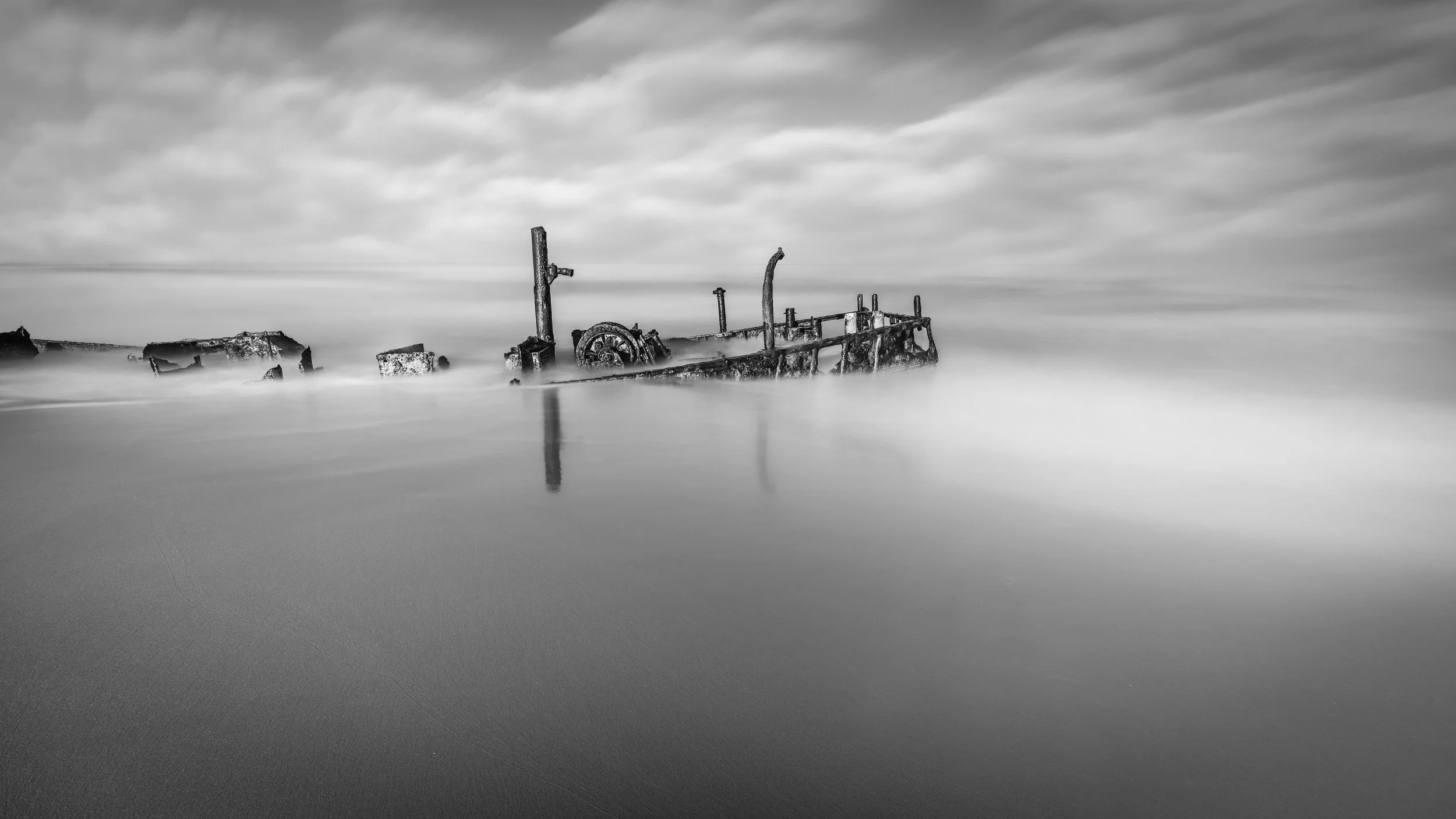 Black and white photo of a submerged rusty shipwreck on a beach with calm water and cloudy sky.