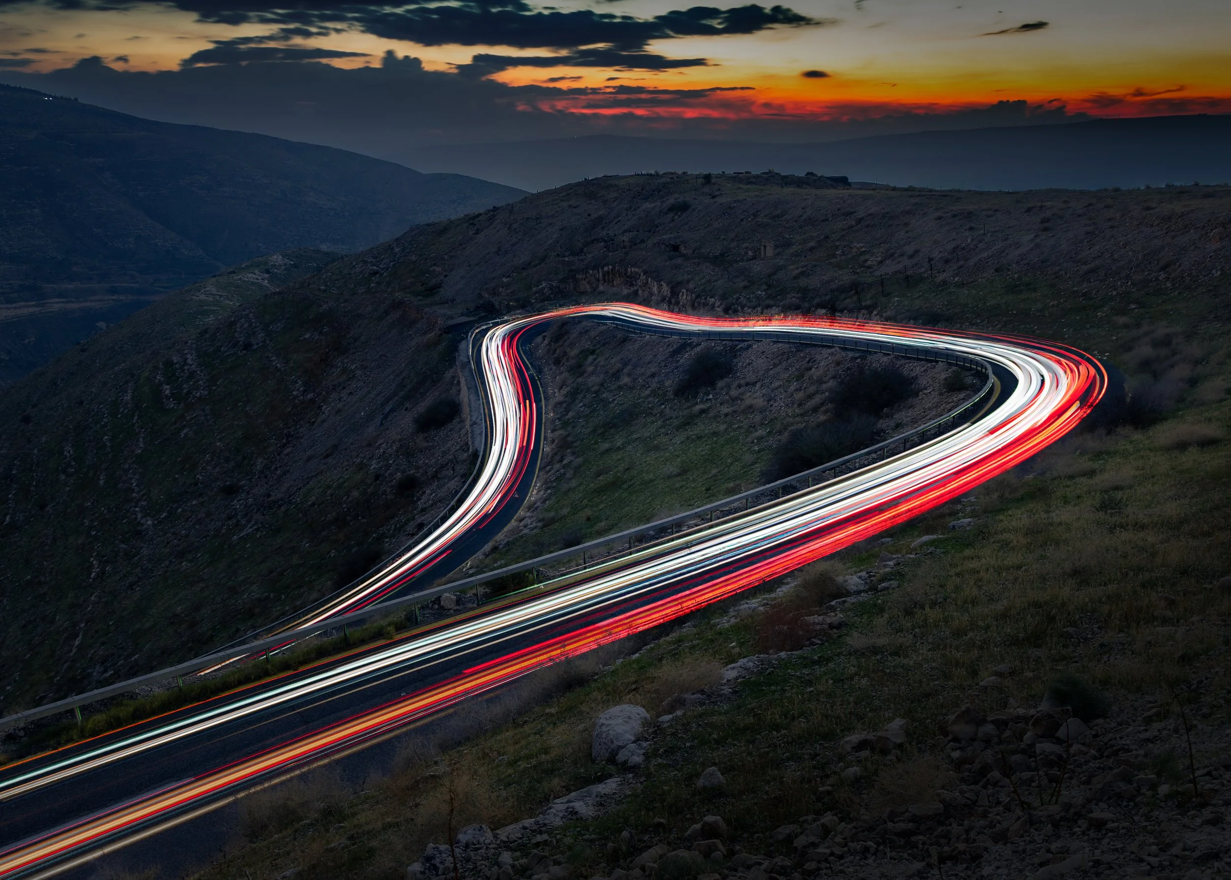 A winding mountain road at dusk with car light trails creating streaks of red and white along the road, surrounded by hills and a sunset sky with clouds.