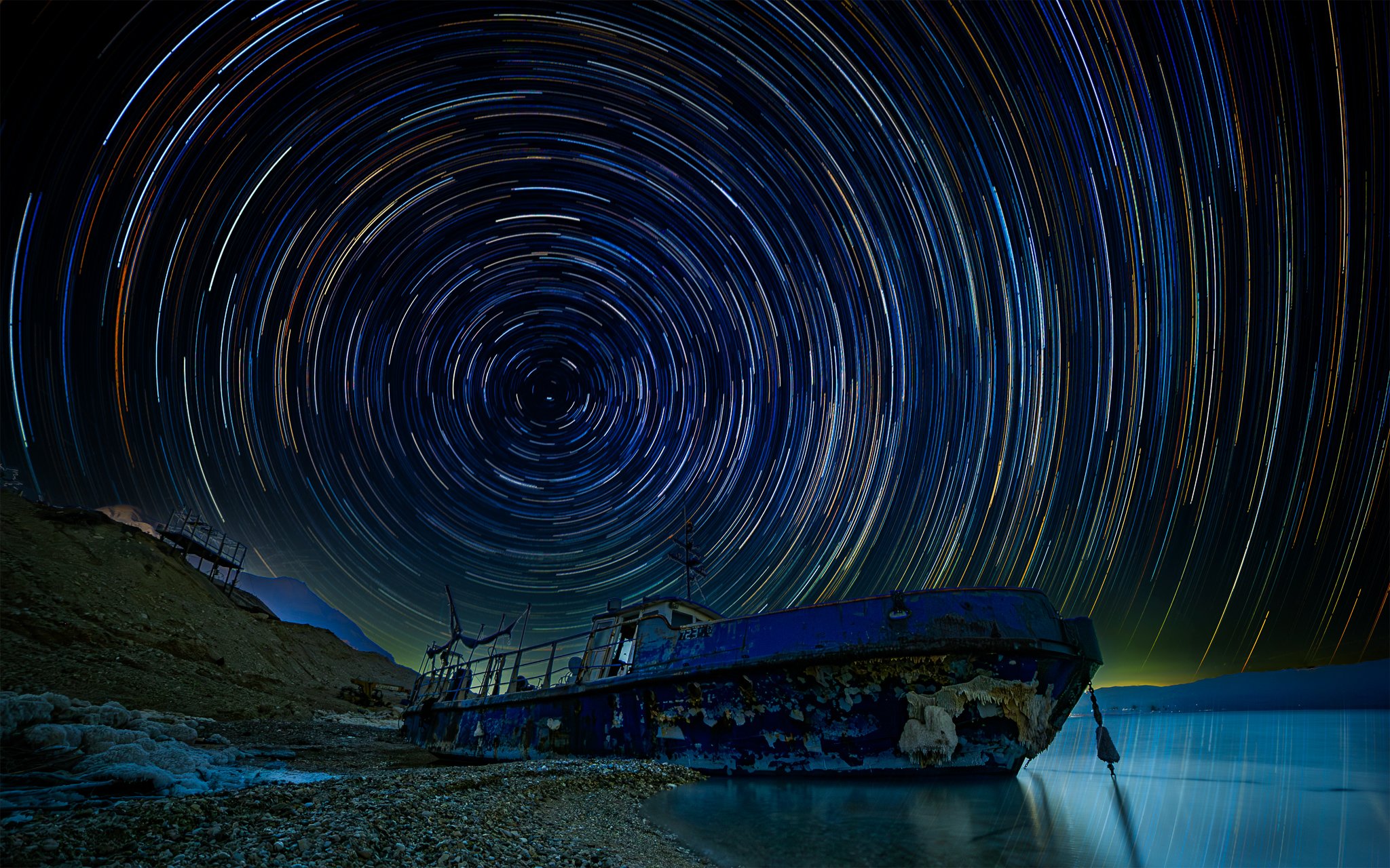 Night sky filled with circular star trail patterns over a landscape with damaged boat and rugged terrain.