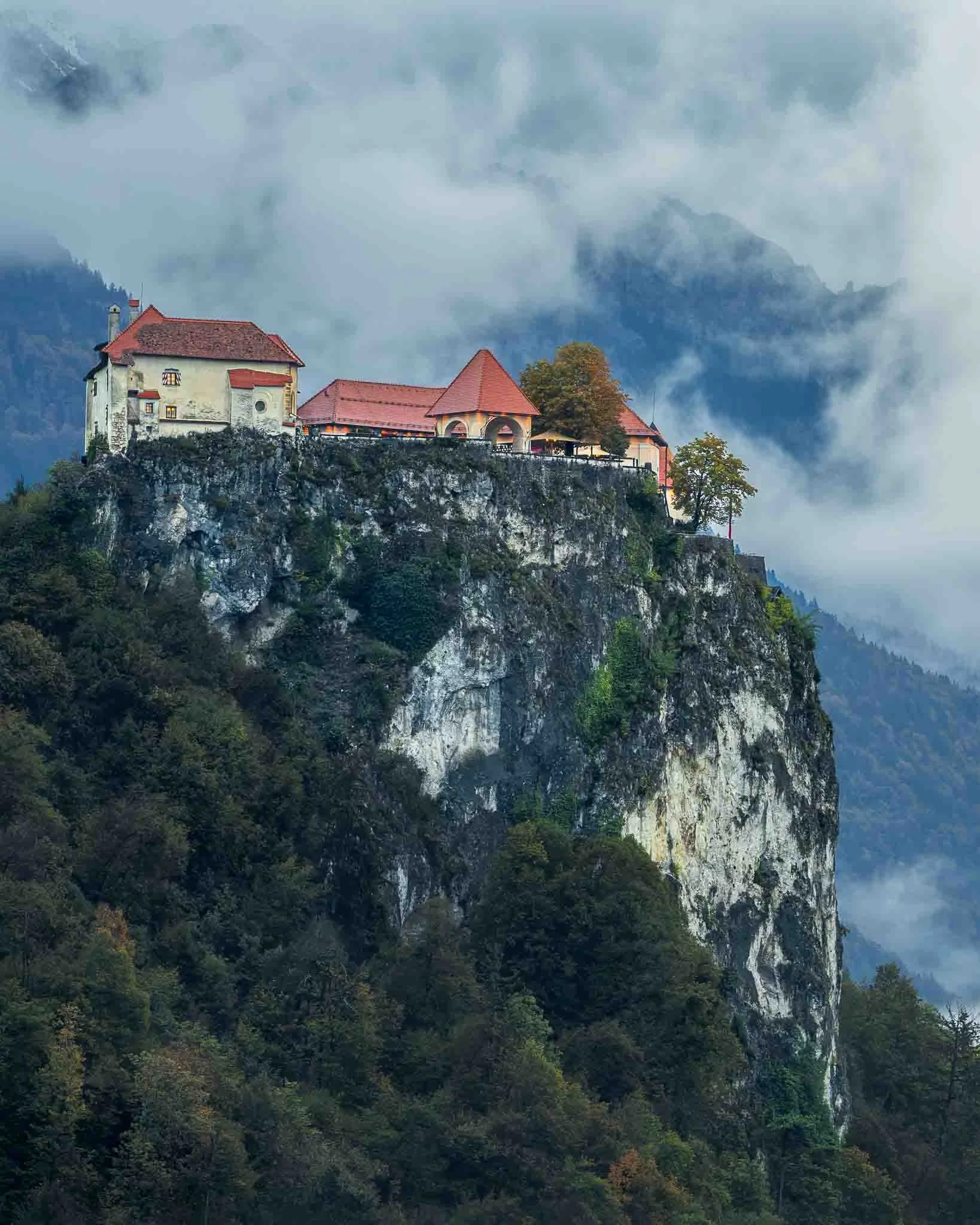 Bled Castle perched on a steep rocky cliff with mist and low clouds drifting across the mountains in Slovenia.