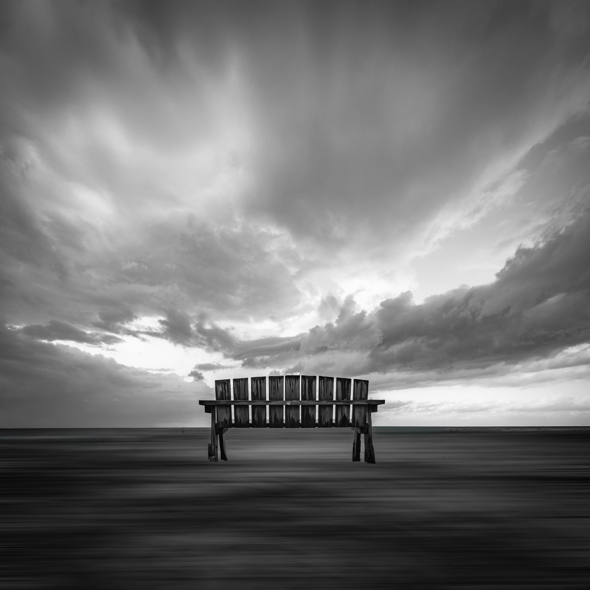 Black and white photo of a lone wooden bench facing an ocean with cloudy sky overhead.