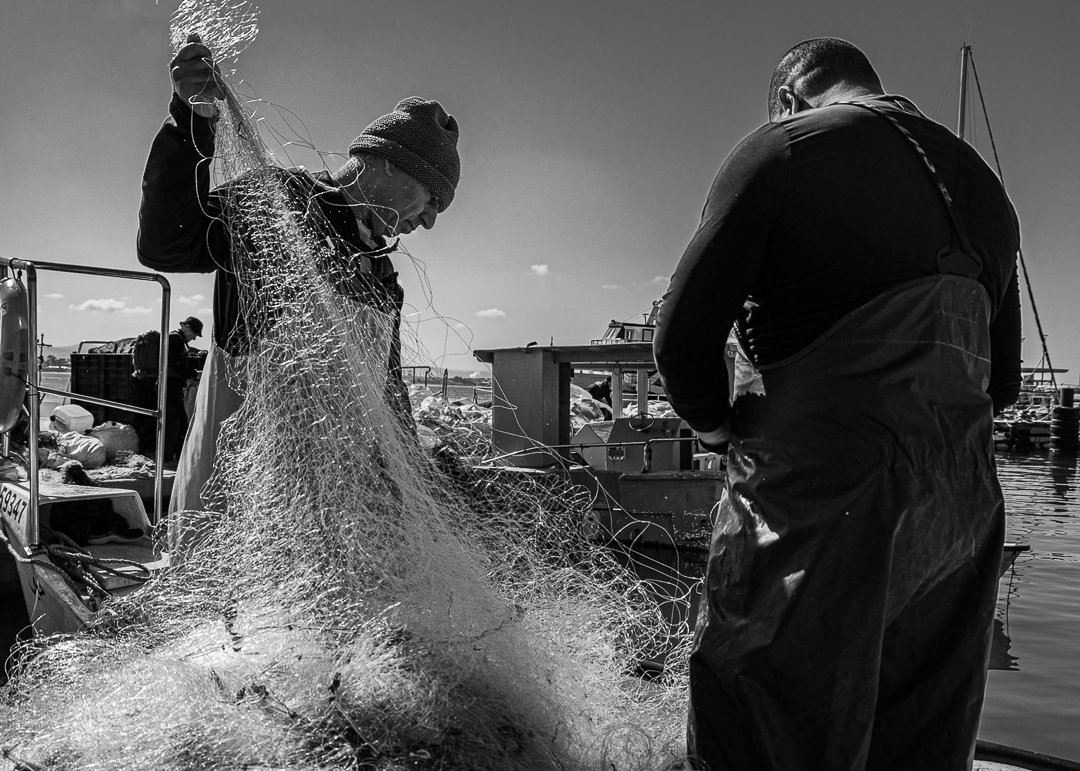 Two fishermen working with fishing nets on a dock near boats in the water, black and white photo.