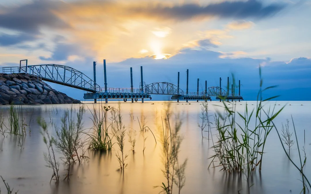 A pier extends into a calm body of water during sunset, with clouds in the sky and grass in the foreground.