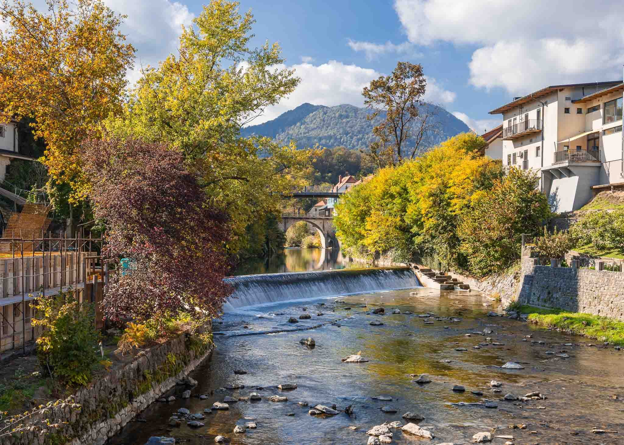 Stone arch bridge over the Selška Sora River in Škofja Loka, framed by autumn trees and riverside buildings, Slovenia.