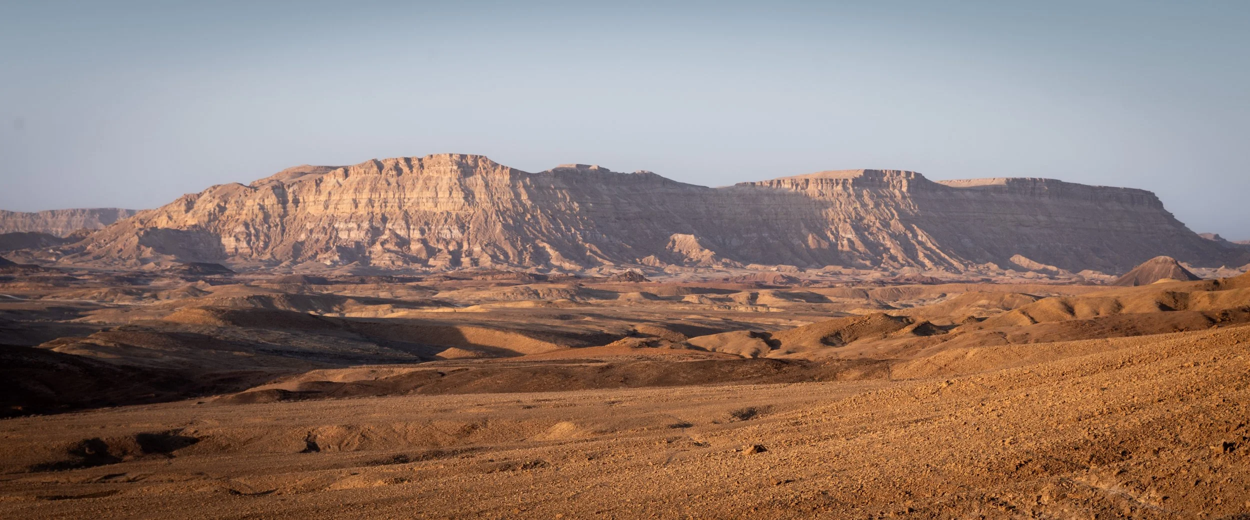 Wide view of a desert landscape with rocky terrain and large plateau mountains in the distance under a clear blue sky.