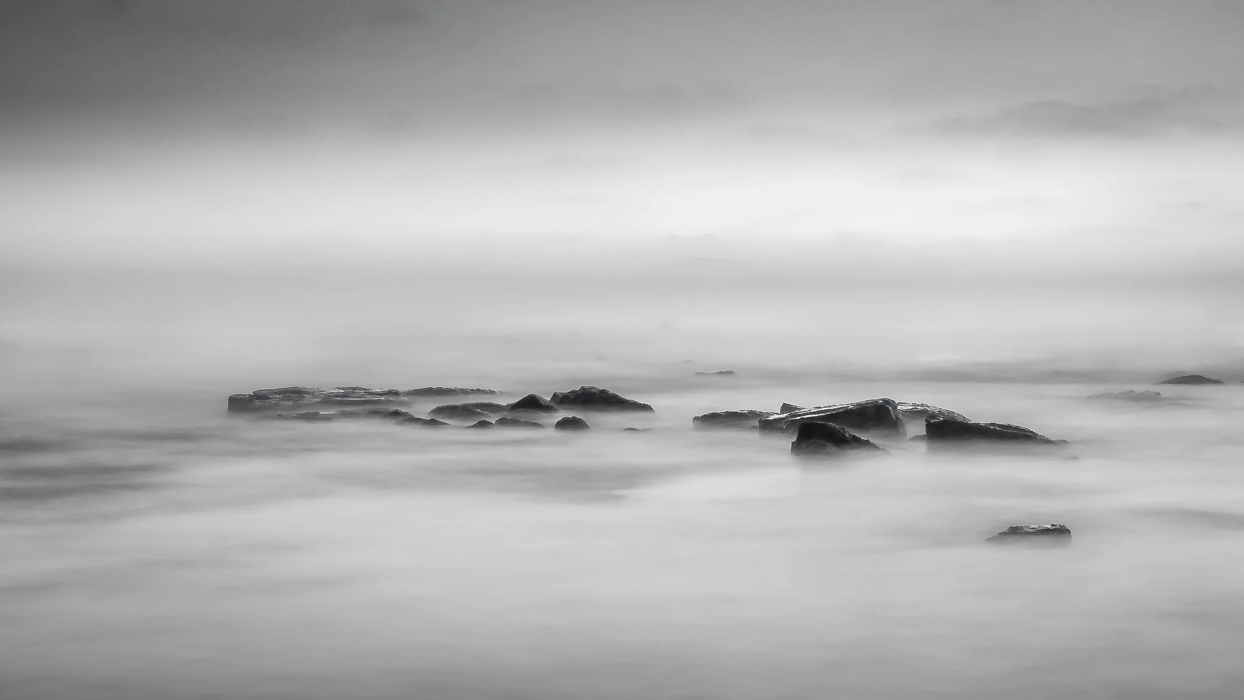 Black and white photo of rocks in calm water with smooth, cloudy sky above.