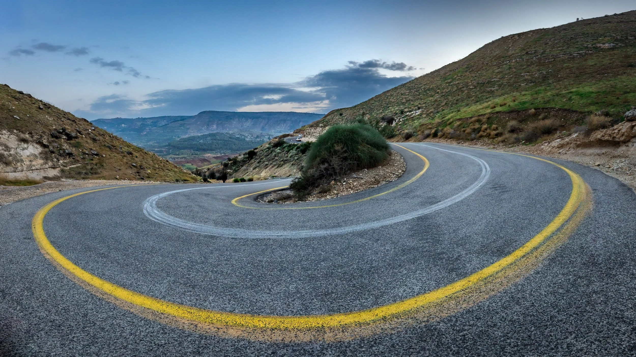 A winding mountain road with yellow dividing lines and a small traffic circle, surrounded by hills and cloudy sky.