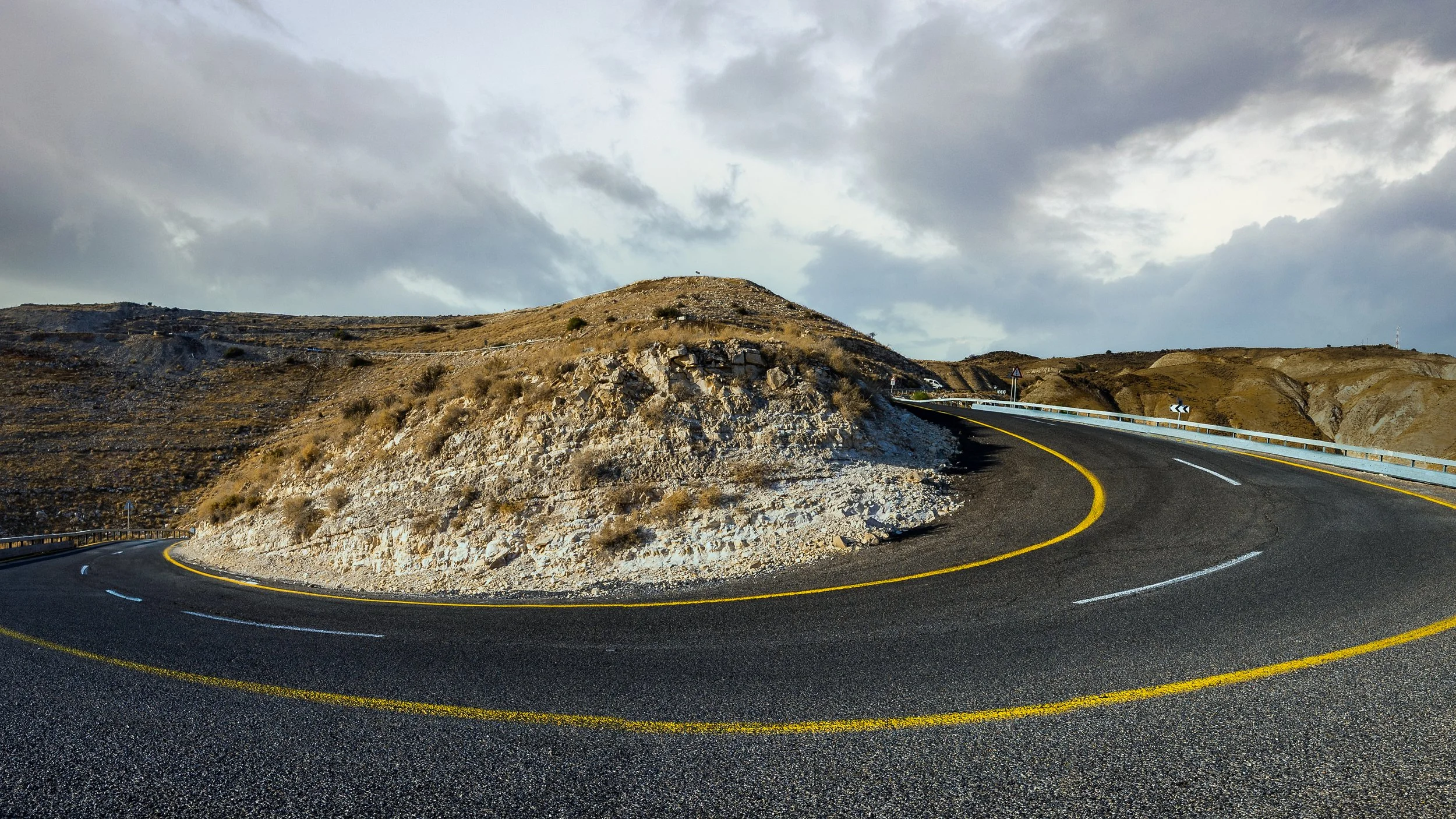 A winding mountain road with black asphalt, yellow and white lane markings, and protective guardrails on each side. Rocky hills and cloudy sky in the background.