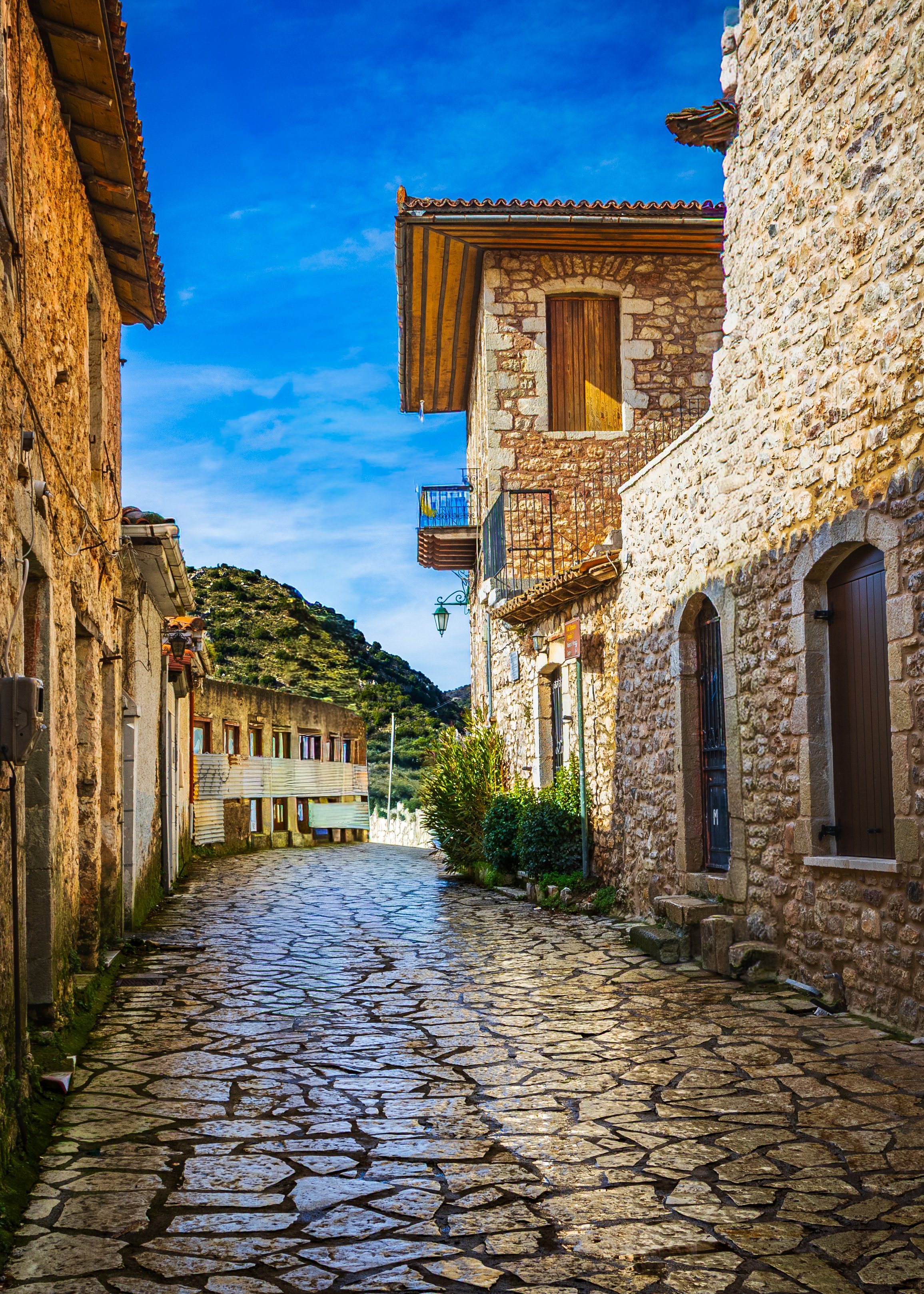 Narrow stone-paved street lined with traditional stone houses under a vivid blue sky.