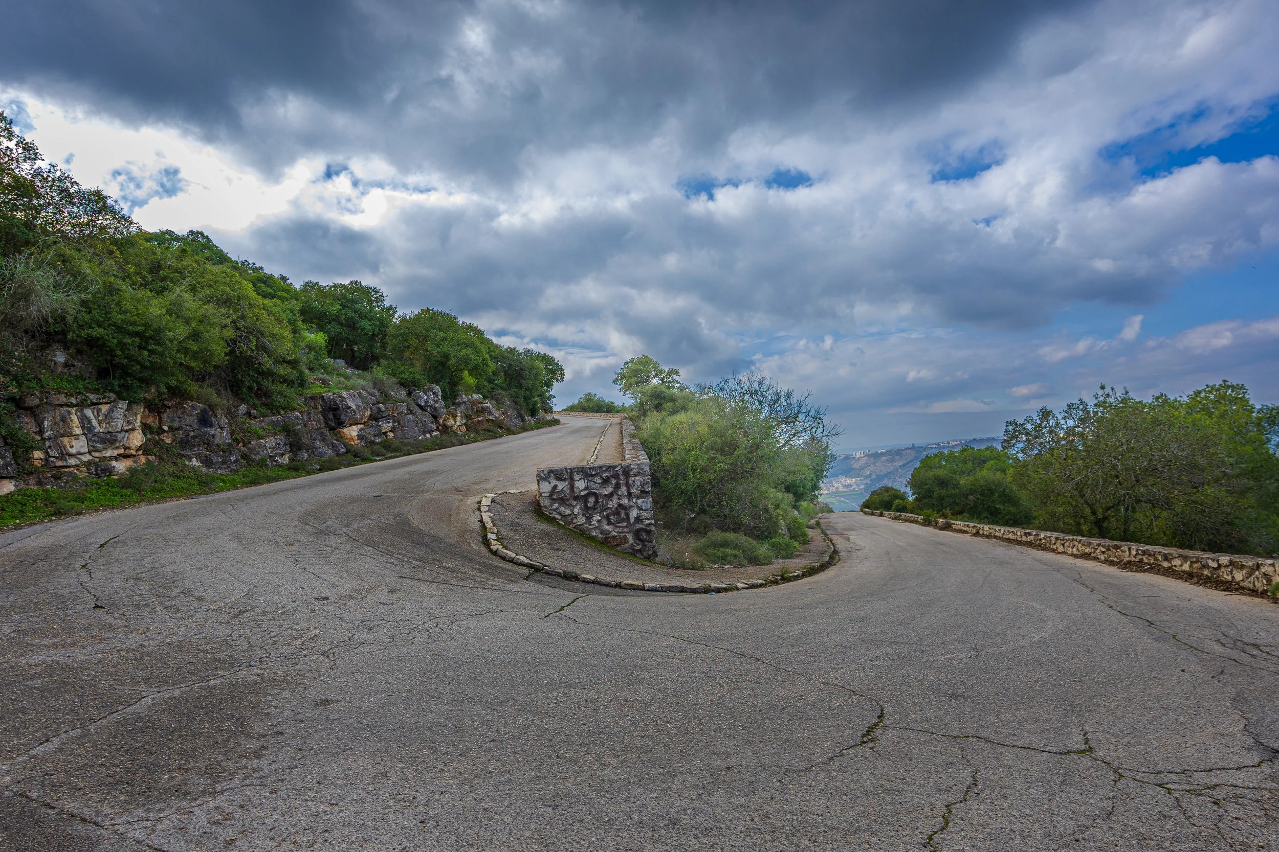 A winding mountain road with cracked pavement, stone guardrails, surrounded by green trees and shrubbery, with a cloudy sky overhead.