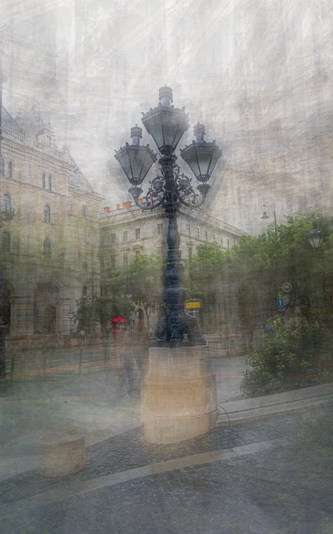 A black ornate vintage street lamp in front of historic European-style buildings, with a foggy or dirty glass surface creating a layered, hazy effect.