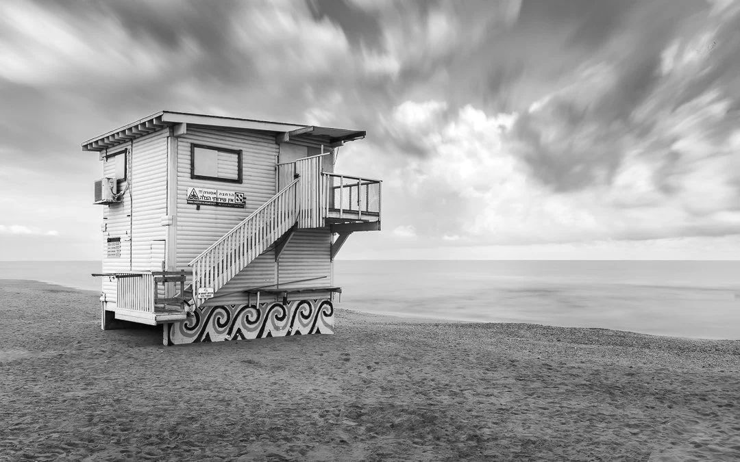 A lifeguard tower on a beach, painted with a wave pattern at the base, under a cloudy sky in black and white.