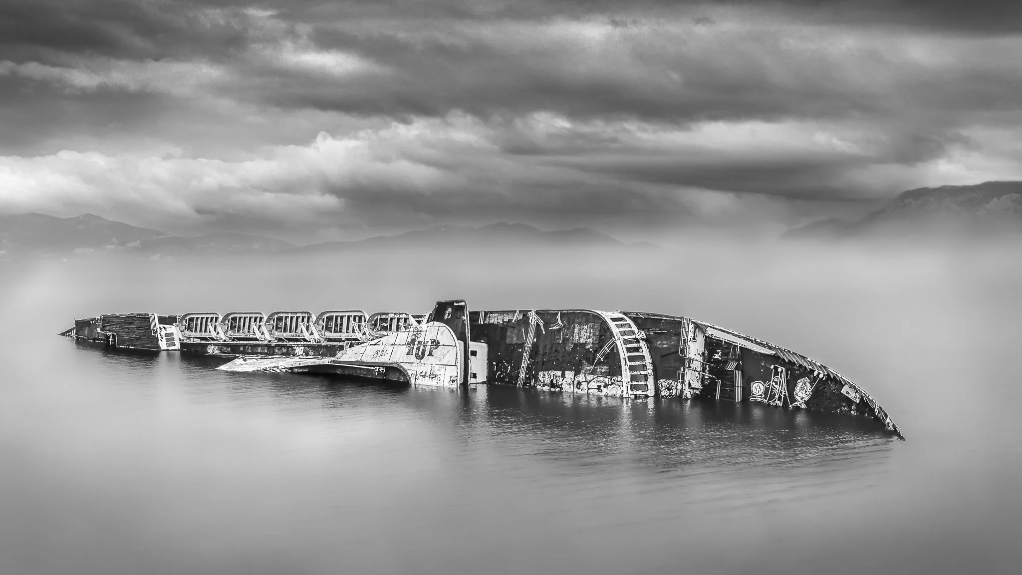 Black and white photo of a sunken shipwreck in calm water with mountains and cloudy sky in the background.