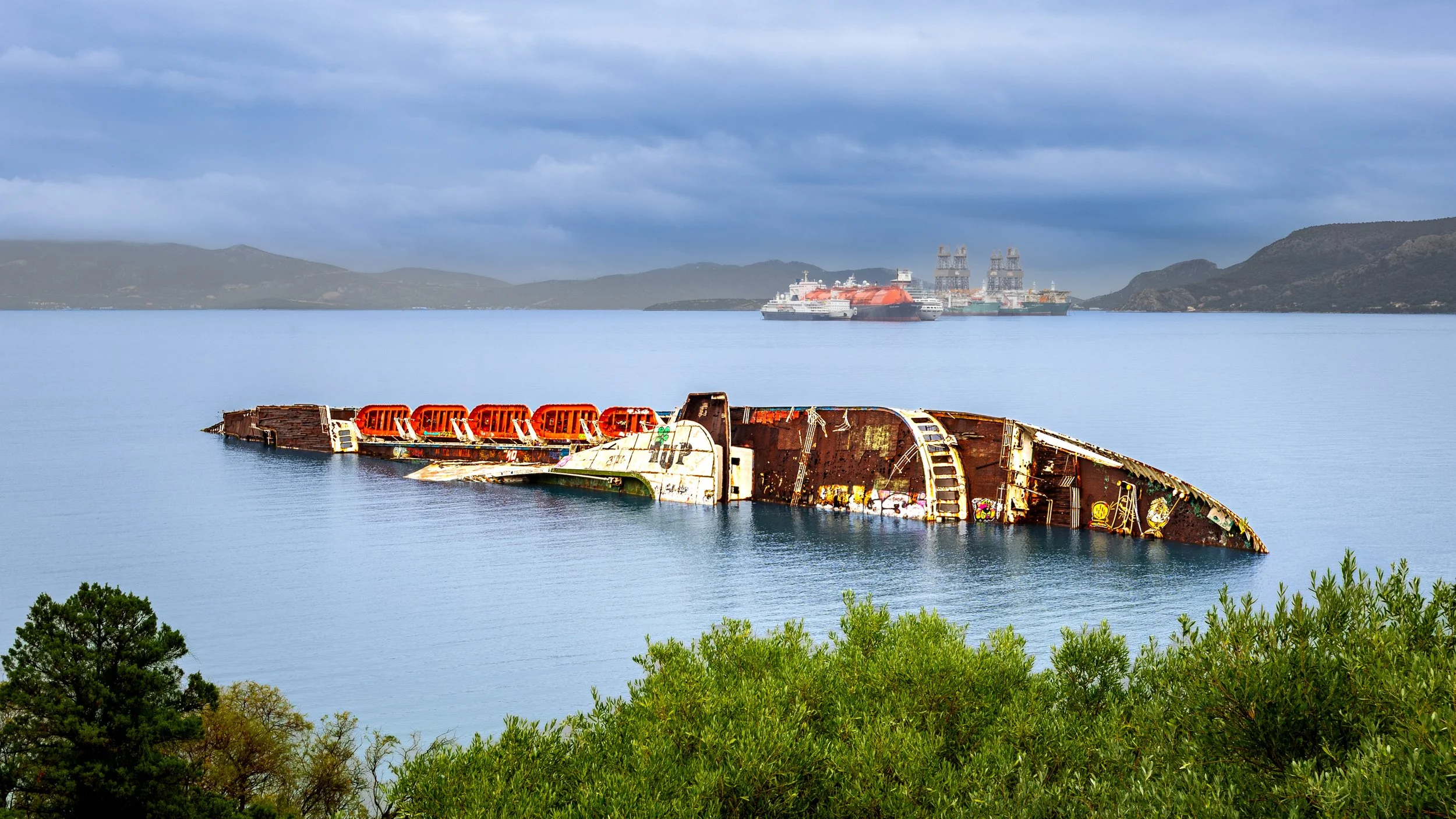 A rusted shipwreck partially submerged in a calm body of water with graffiti, greenery in the foreground, and a large oil tanker in the background under cloudy skies  photography by erez nudmanov