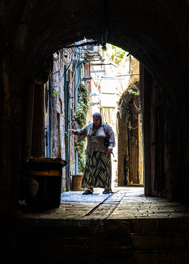 A woman with a headscarf standing in a narrow, dark alleyway with stone walls, looking at or touching items on the wall.