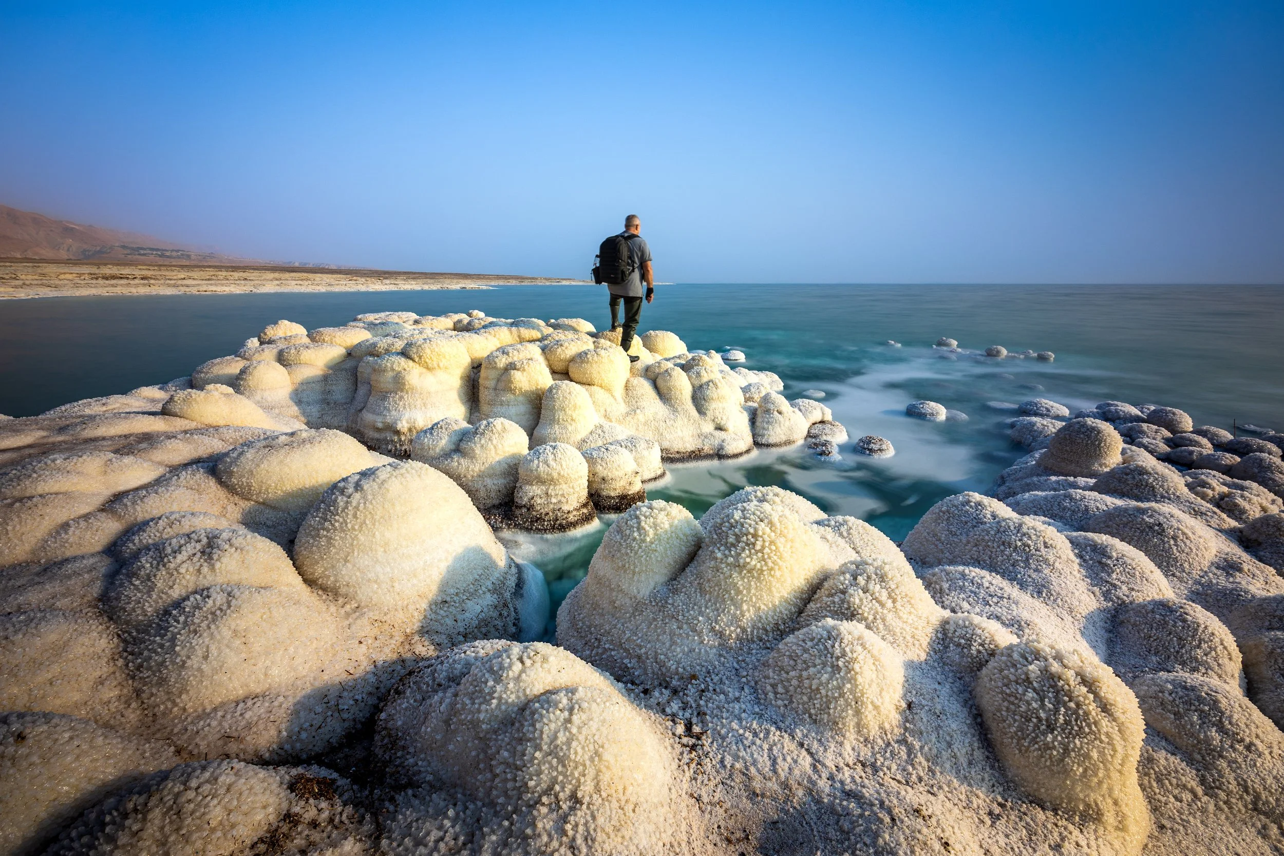 A person standing on salt formations in a desert-like area beside the ocean, with a distant coastline and clear blue sky.