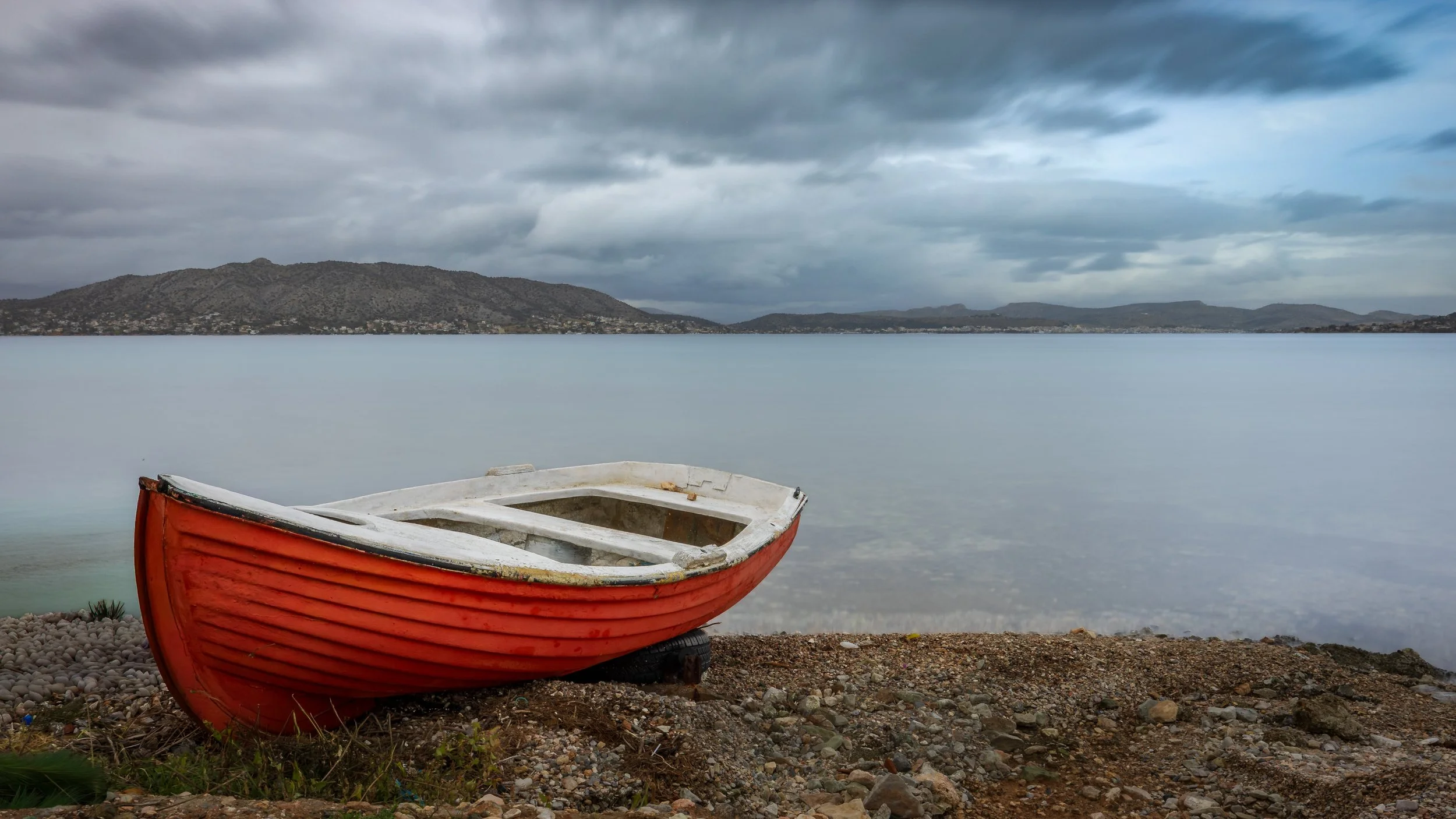 A red and white boat resting on a rocky beach with calm water and mountains under a cloudy sky in the background, photograph by erez nudmanov