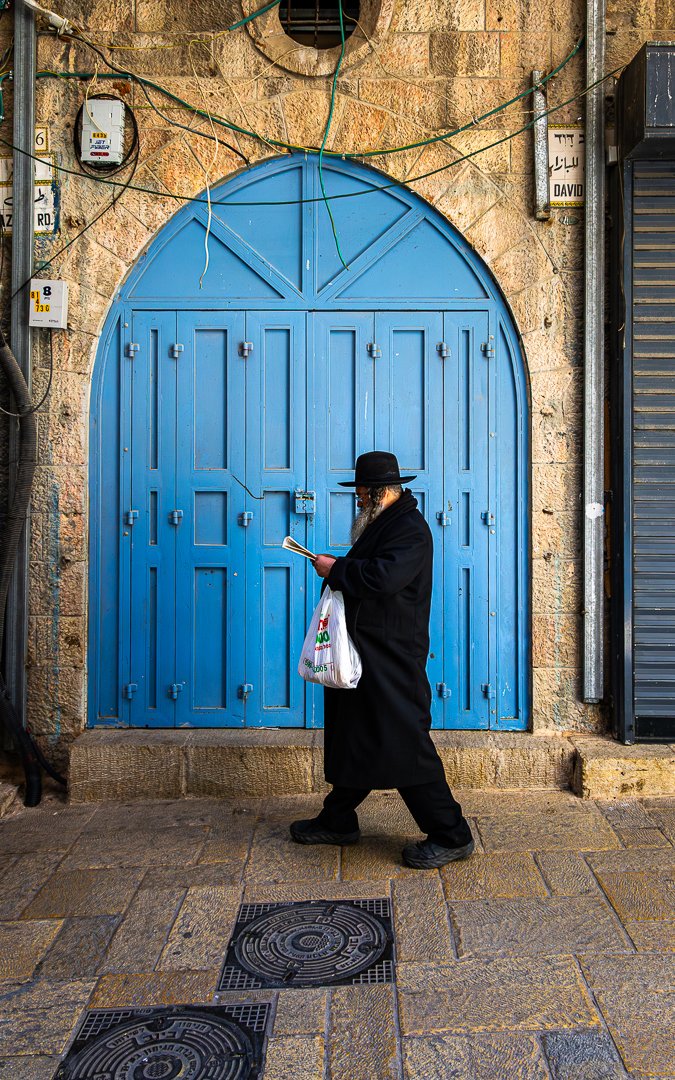 An Orthodox Jewish man with a black hat and long black coat reading a book while walking past a blue arched door on a stone building in an urban setting.