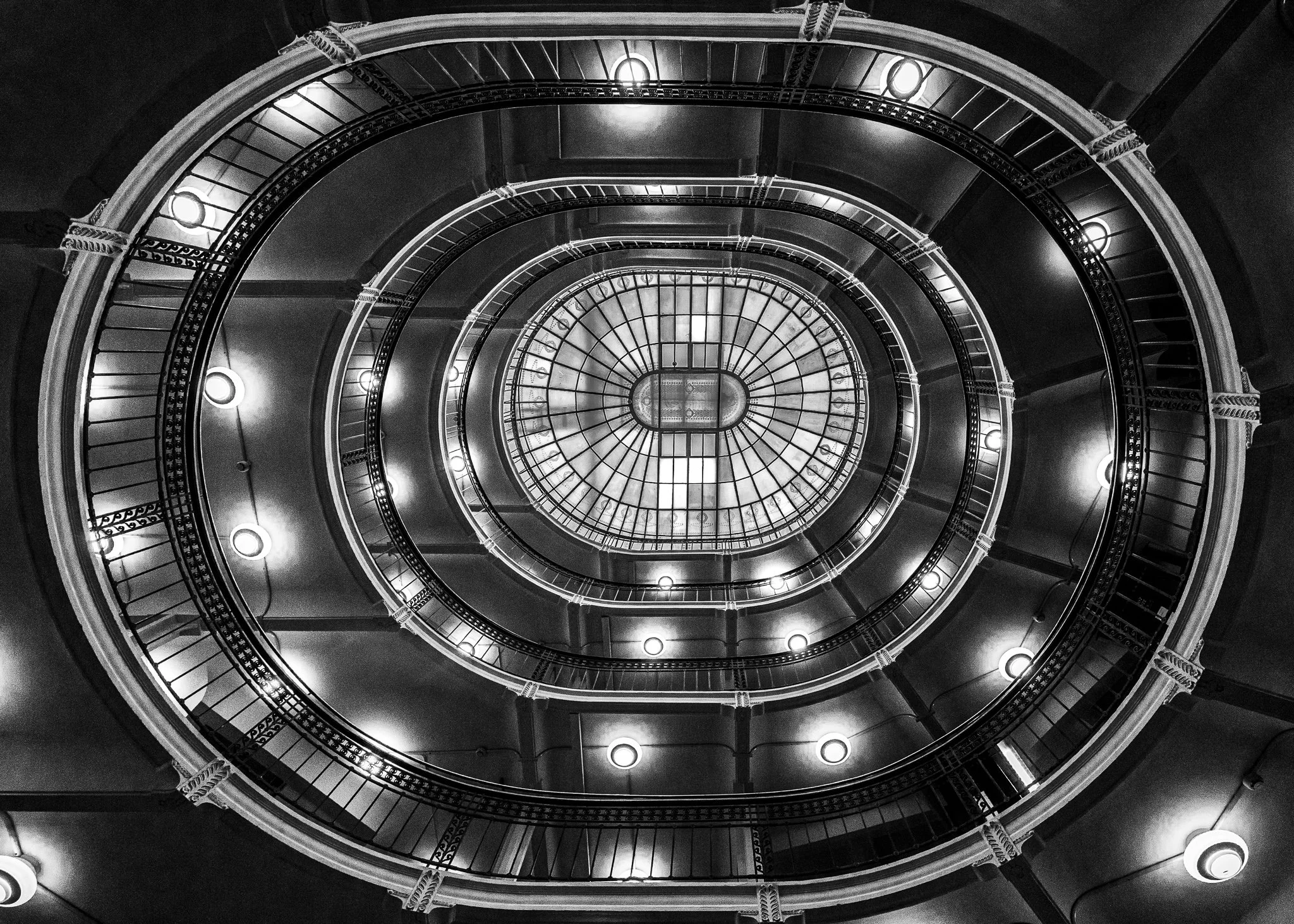 Black and white photo looking up a spiral staircase with ornate railings, circular lights, and a glass ceiling at the top.