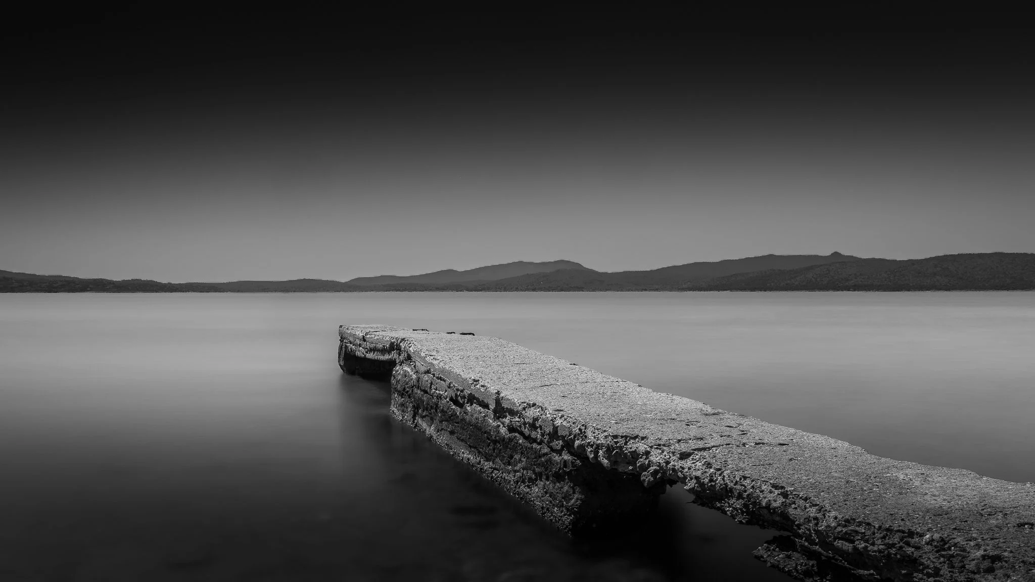 A black-and-white photograph of a stone pier extending into a calm body of water with distant mountains and a clear sky in the background.