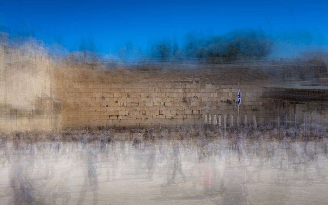 Blurred photo of a historical stone wall with a Greek flag, outdoor seating, and umbrellas in front, under a clear blue sky.