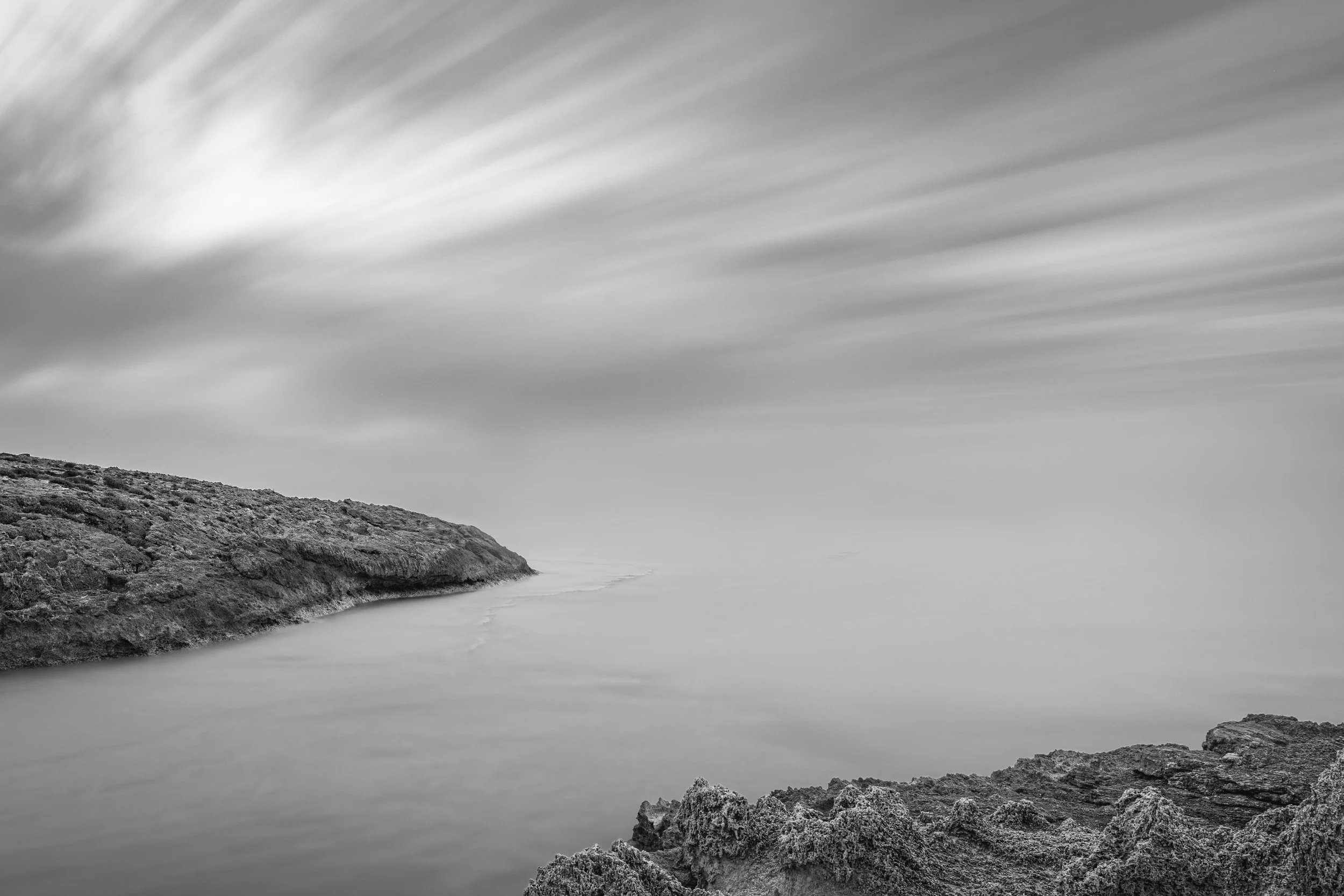Black and white long exposure photo of a rocky coastal landscape with a calm body of water and a sky with streaked clouds.