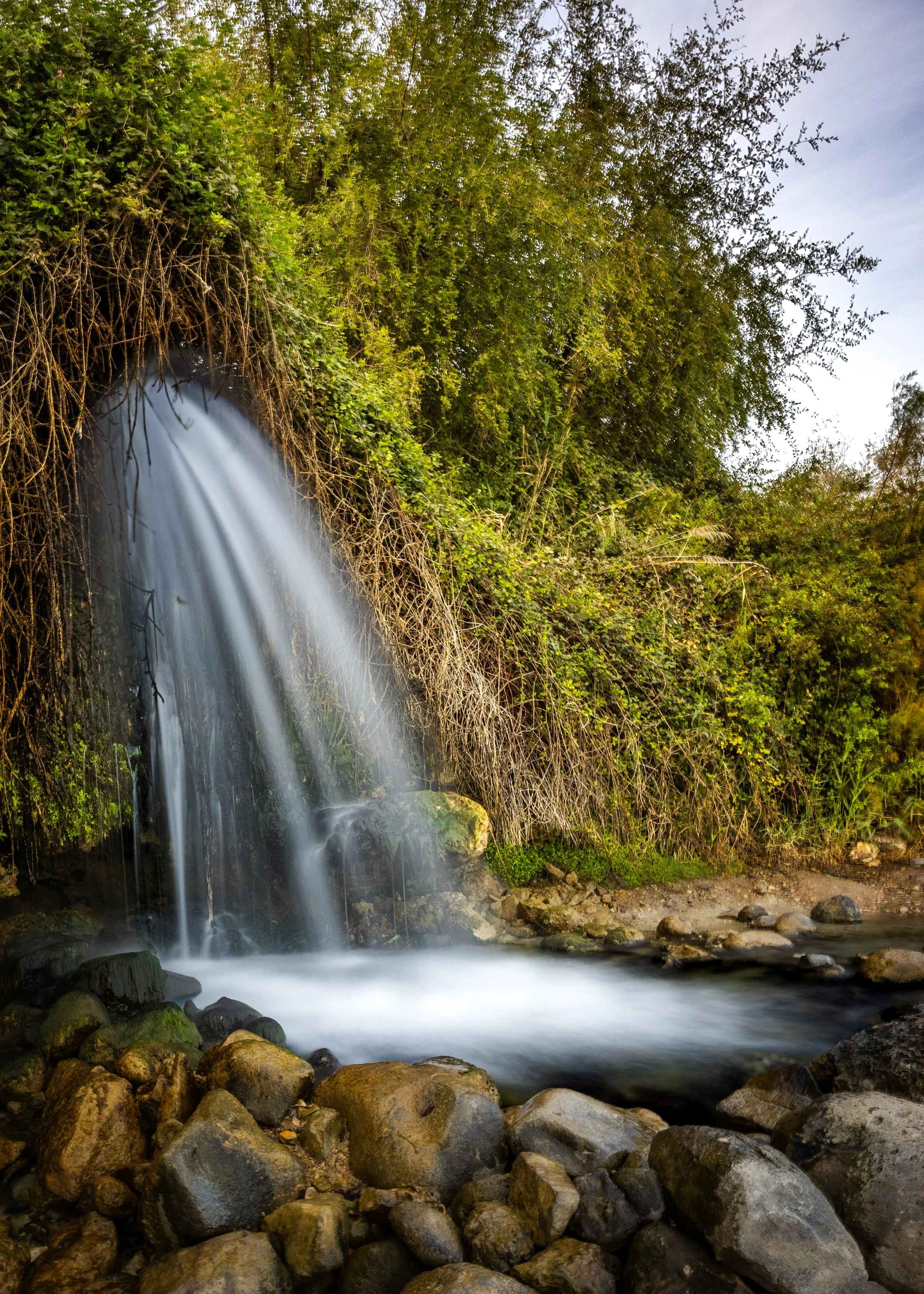 Small waterfall flowing over rocks into a stream, surrounded by dense green foliage photography by erez nudmanov