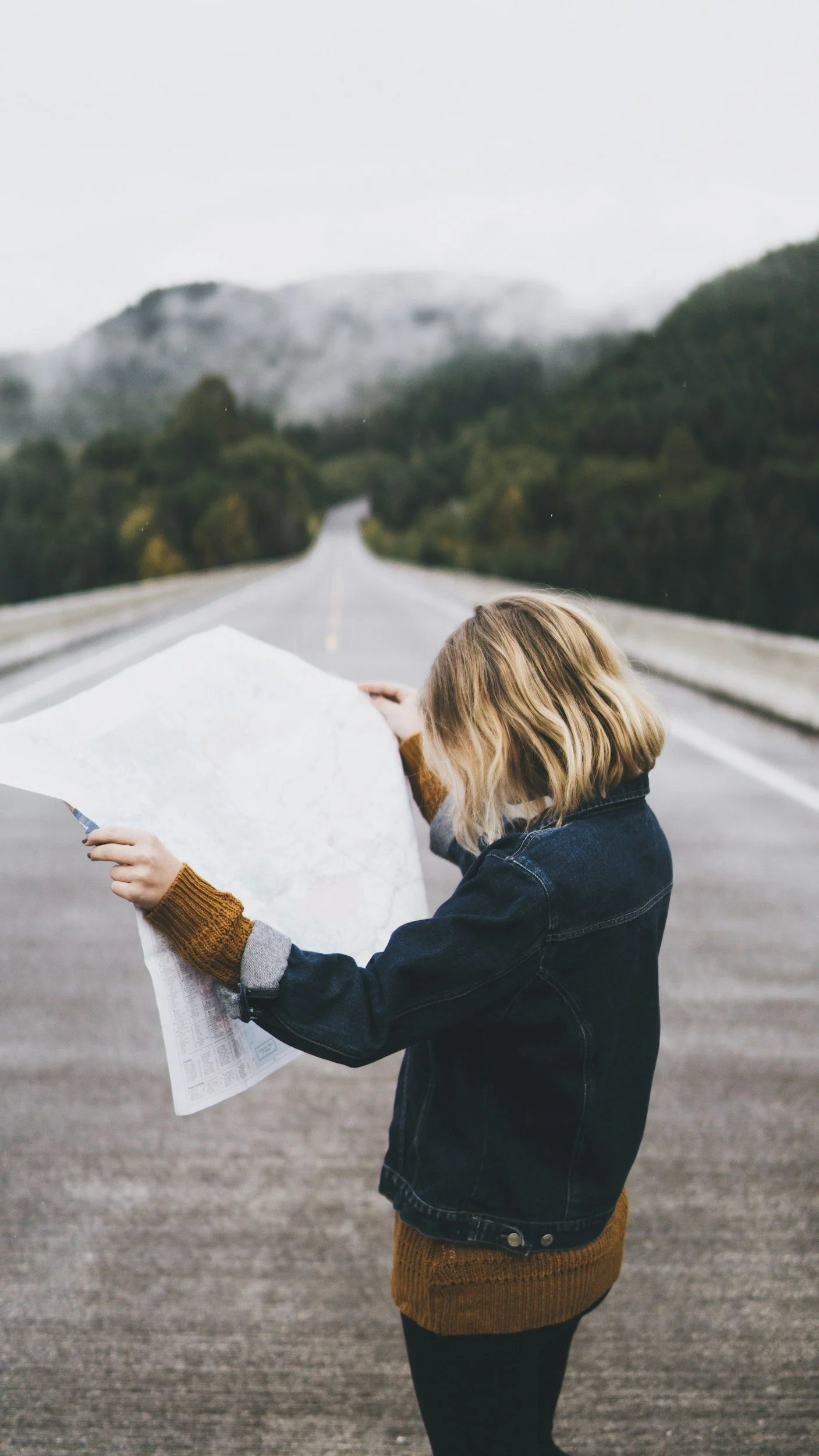 A person with shoulder-length blonde hair, wearing a dark jacket and orange-brown sweater, stands on a deserted road holding a large map, with scenic hills and trees in the background.