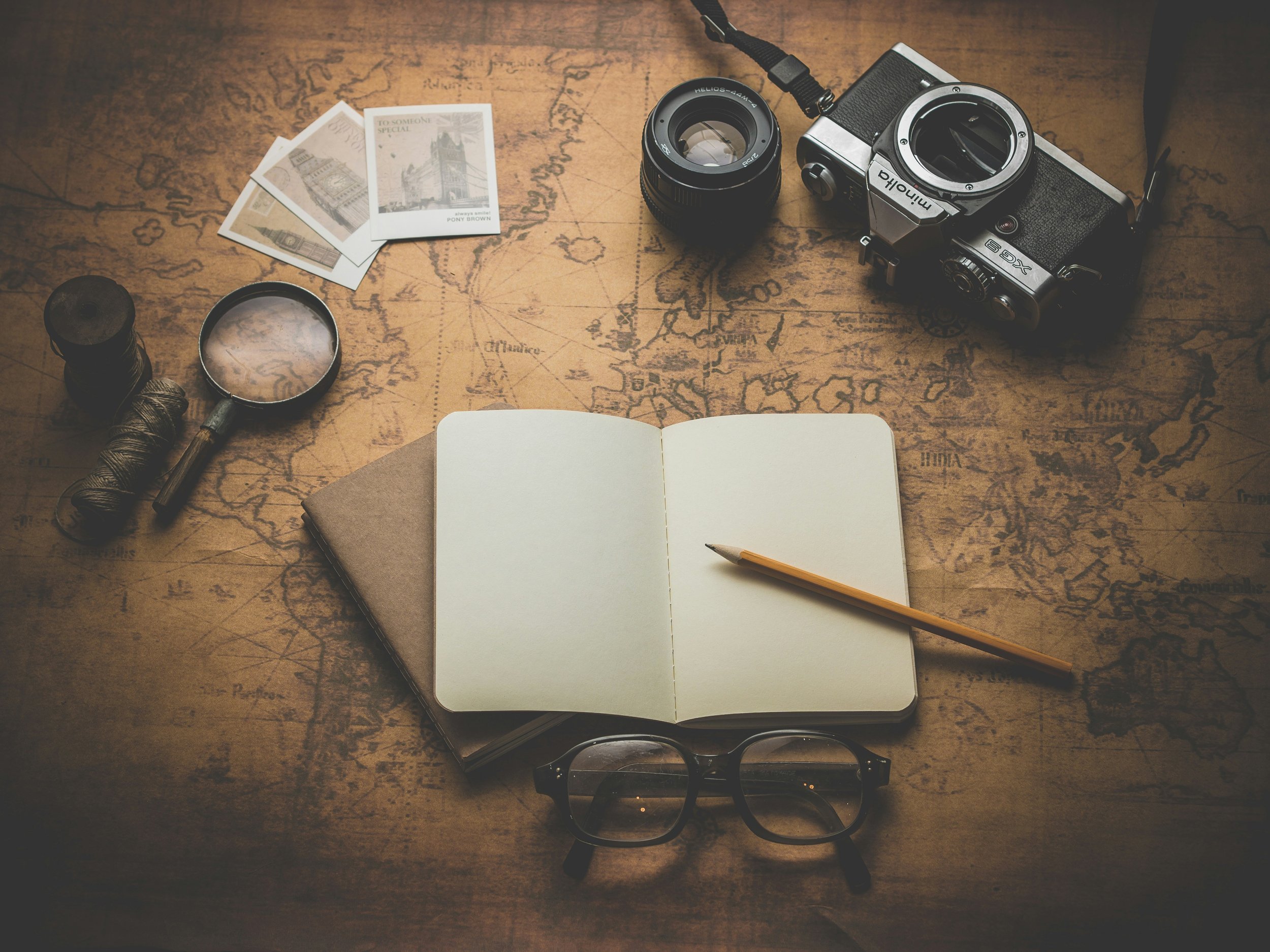 Travel or exploration setup with a vintage map, a camera, a jar of string, a magnifying glass, postcards, a notebook with a pencil, glasses, and small containers on a wooden surface.