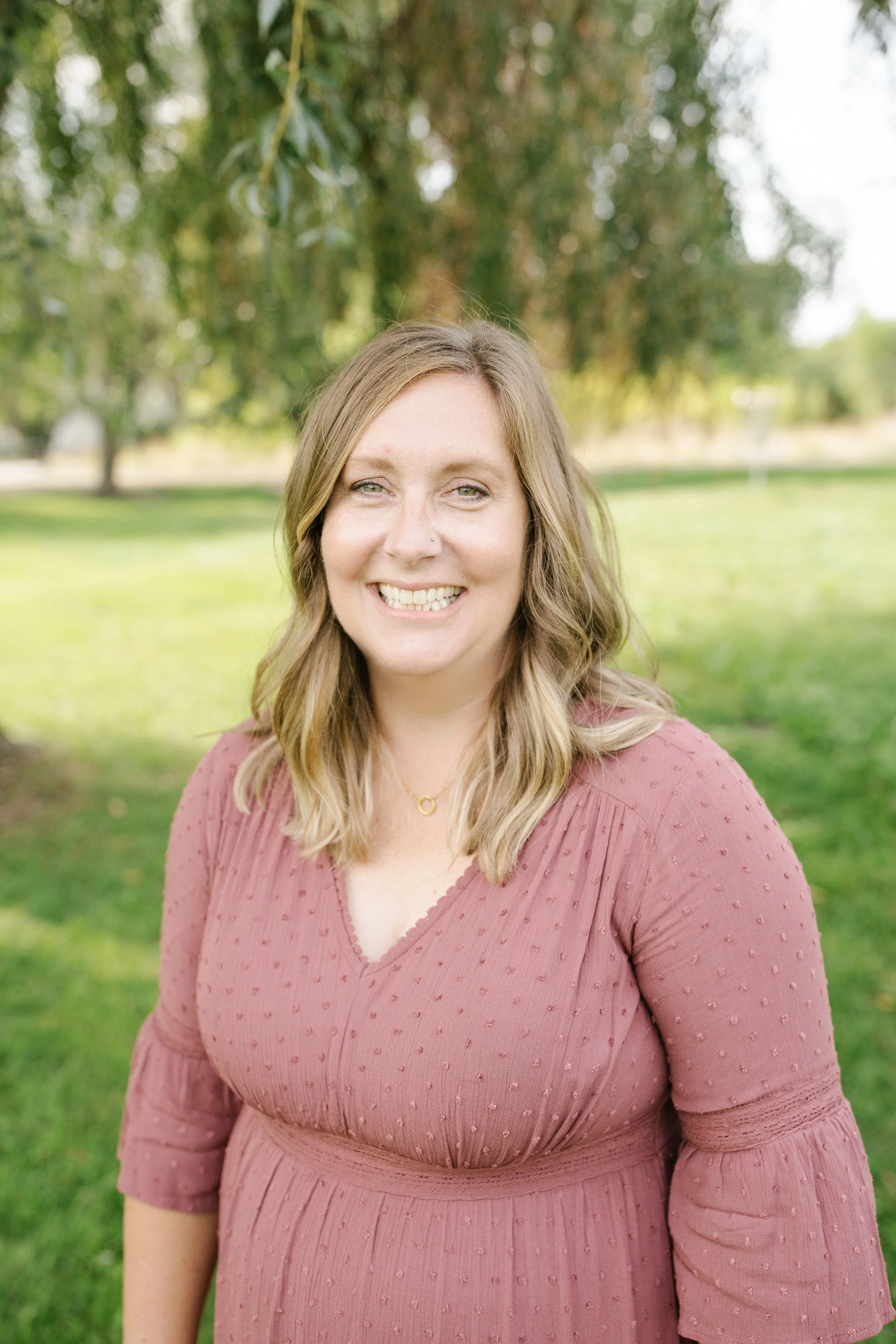 A woman with shoulder-length blonde hair smiling outdoors in a park, wearing a mauve dress with textured dots.