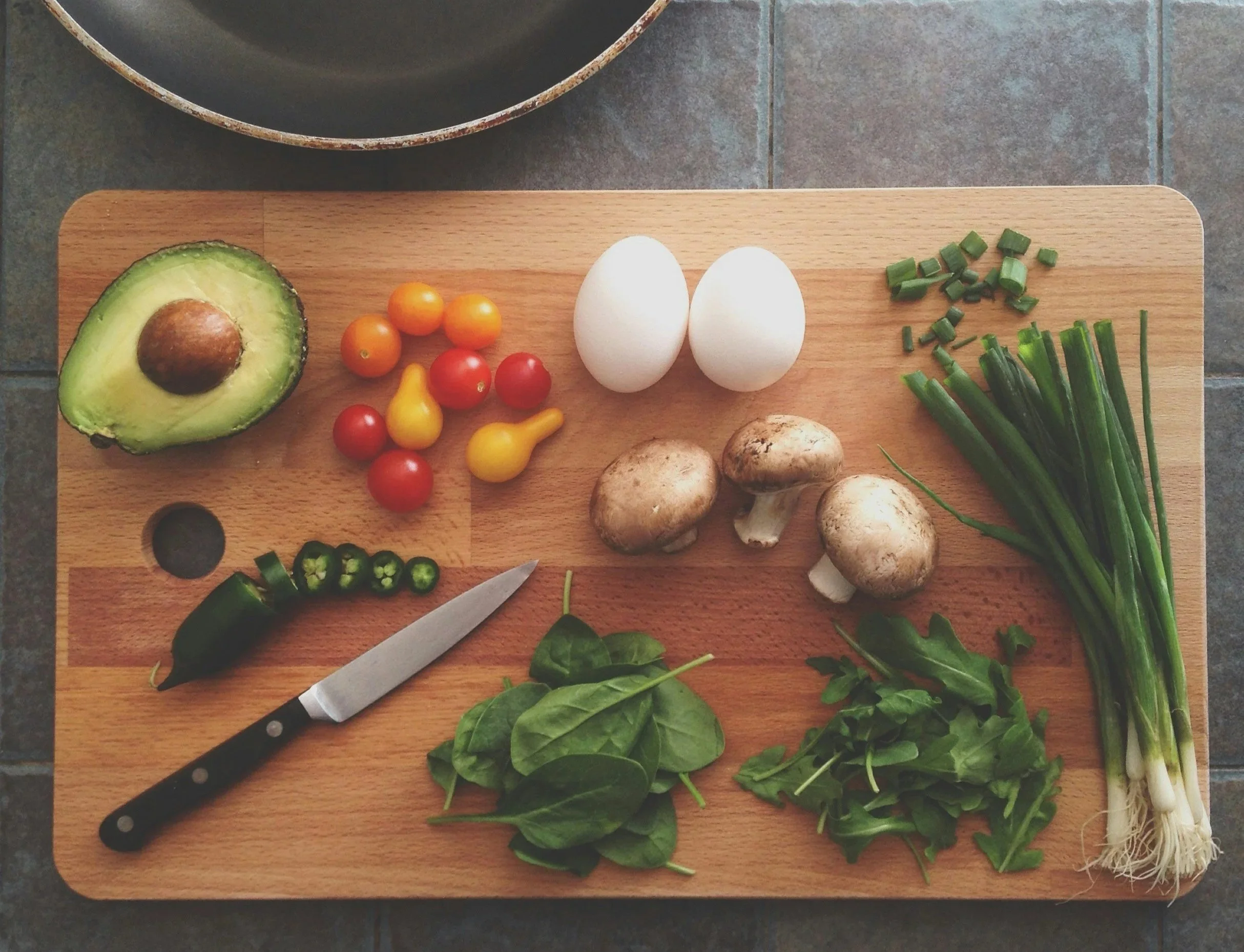 Top-down view of a wooden cutting board with various vegetables including avocado, cherry tomatoes, mushrooms, green onions, and spinach, with a chef's knife.