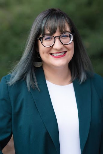 A woman with glasses and shoulder-length dark hair outdoors, wearing a teal blazer and white top.