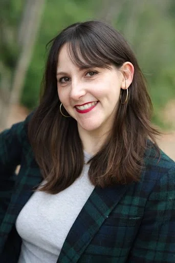 A woman with shoulder-length brown hair, wearing hoop earrings, a gray shirt, and a dark plaid jacket, smiling outdoors with a blurred green background.