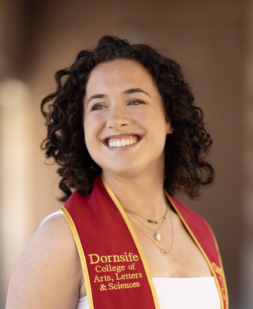 A woman with curly brown hair, wearing a white top and a red stole with yellow trim that reads 'Dornsife College of Arts, Letters & Sciences.' She is smiling and looking to the side, with a blurred background.